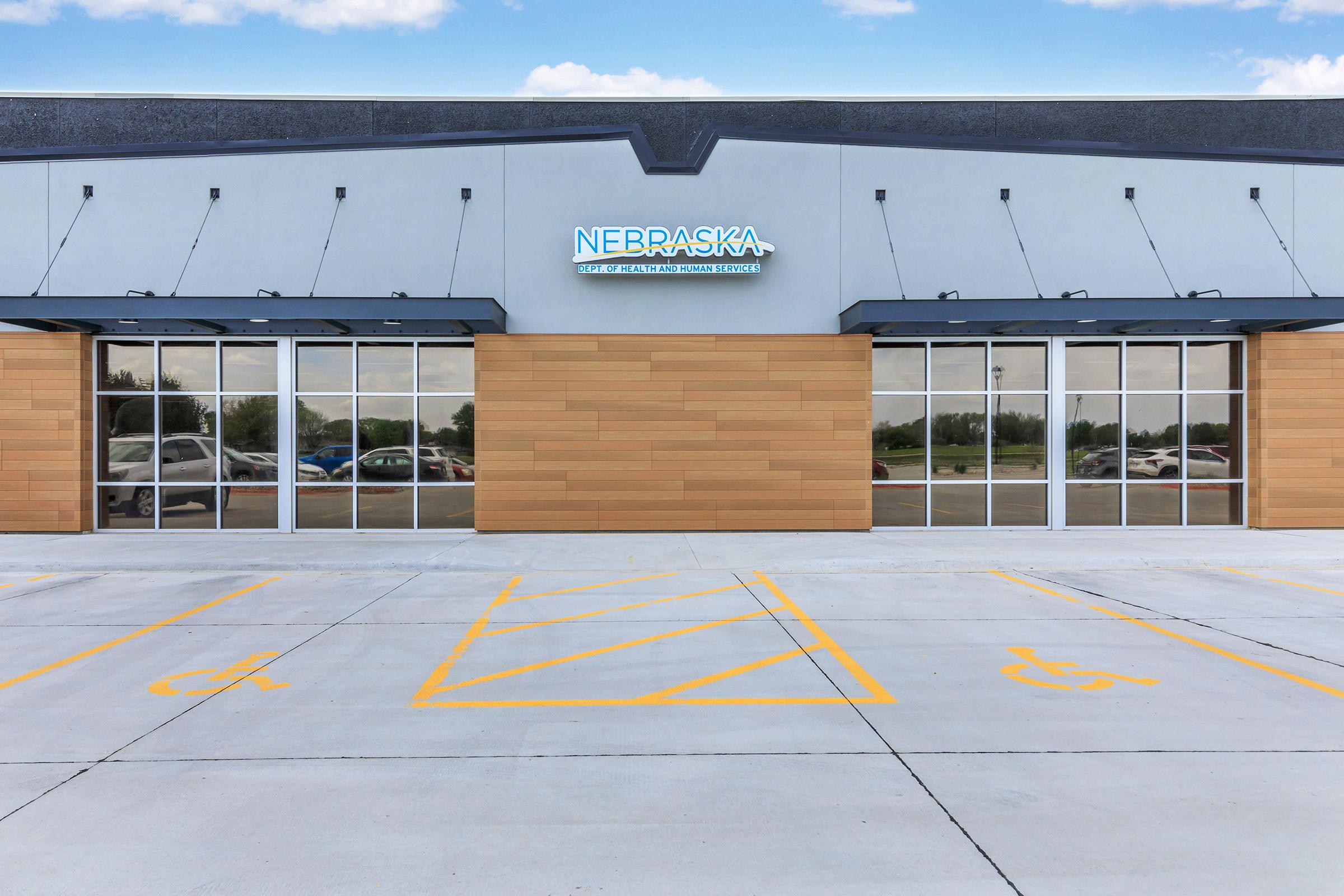 Facade of a modern building featuring large glass windows and a wooden panel accent, with a sign reading "NEBRASKA" illuminated above the entrance. The foreground shows a parking area with clearly marked accessible parking spaces. The sky is partly cloudy.