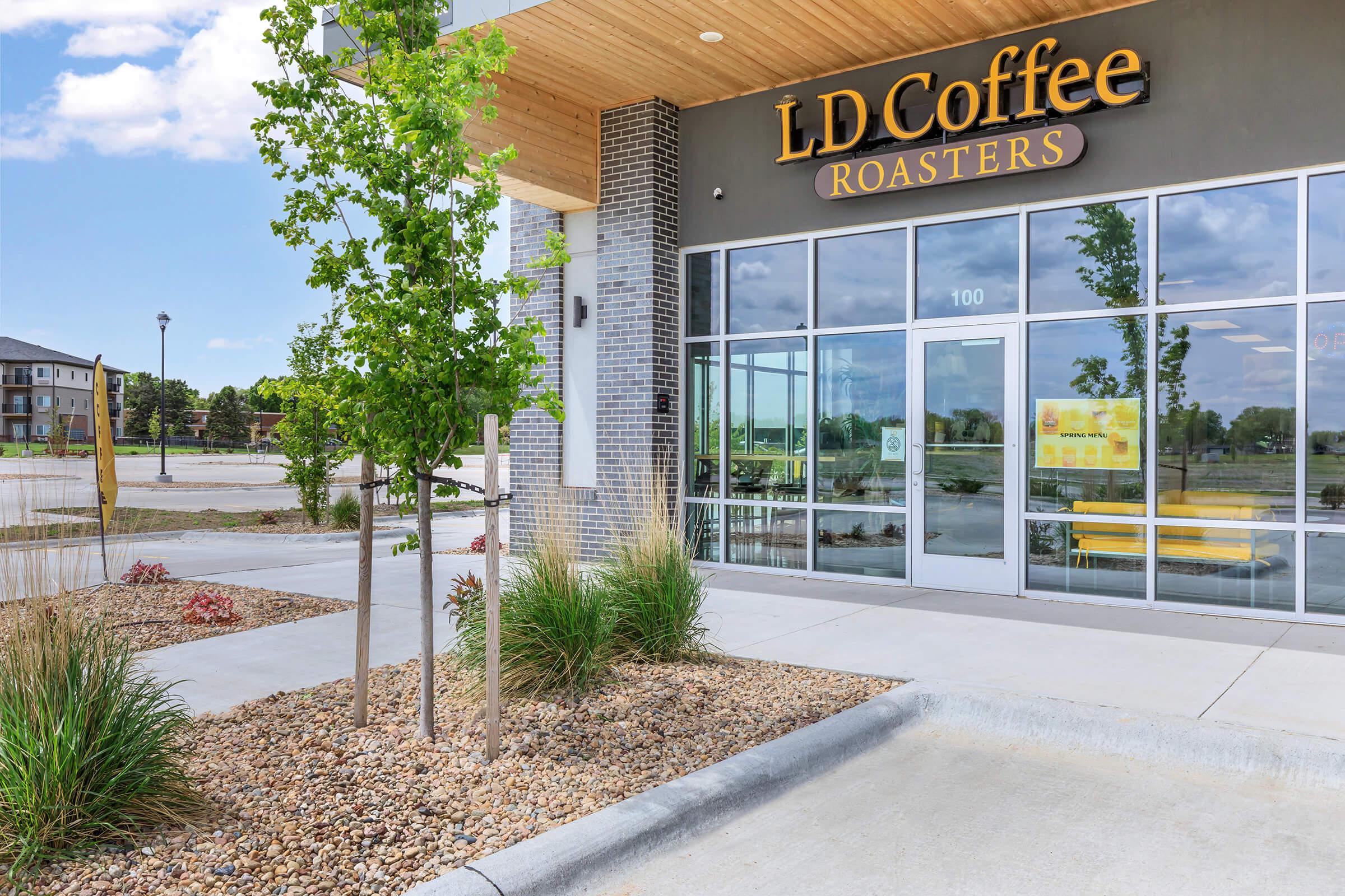 Exterior view of LD Coffee Roasters with a modern design, featuring large glass windows, a wooden overhang, and a landscaped area with small trees and decorative stones. The storefront is inviting, with visible signage above the entrance. A spacious area is seen in the background.