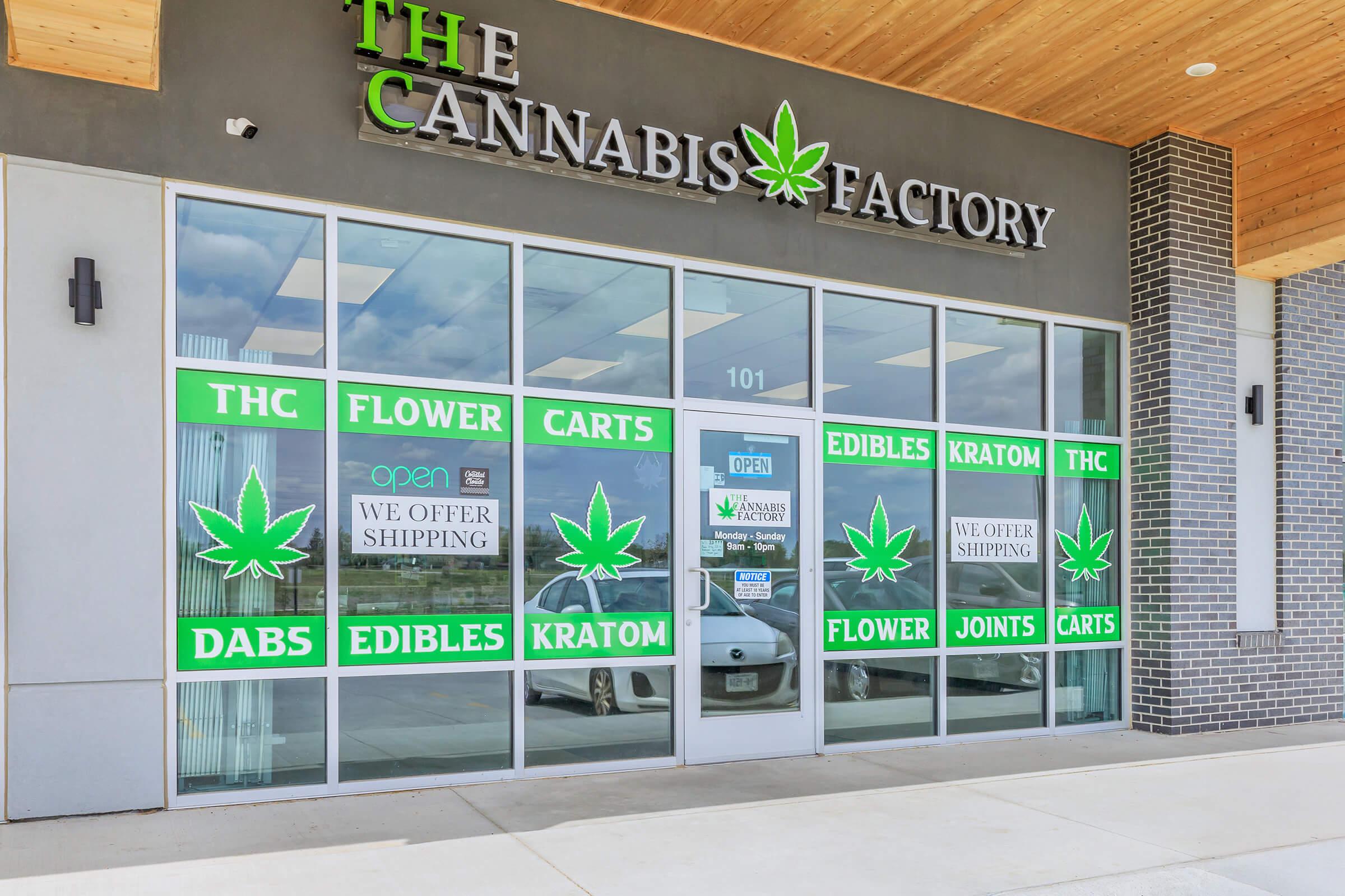 Storefront of "The Cannabis Factory," featuring large green cannabis leaf graphics and advertisements for various products including THC, flower, edibles, carts, dabs, and kratom. The entrance displays an "Open" sign and includes shipping information. The building has a modern design with a wooden overhang.