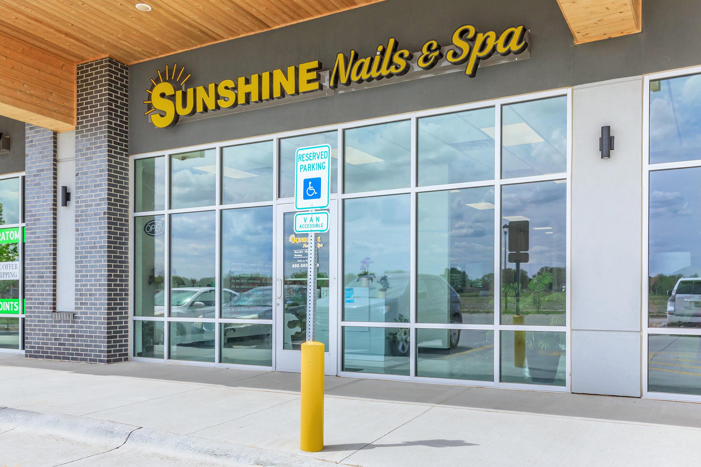 Exterior view of Sunshine Nails & Spa, featuring large glass windows and a modern design. The sign is prominently displayed above the entrance, and there is a reserved parking sign for disabled access in front of the building. The sky is partly cloudy, adding to the inviting atmosphere.