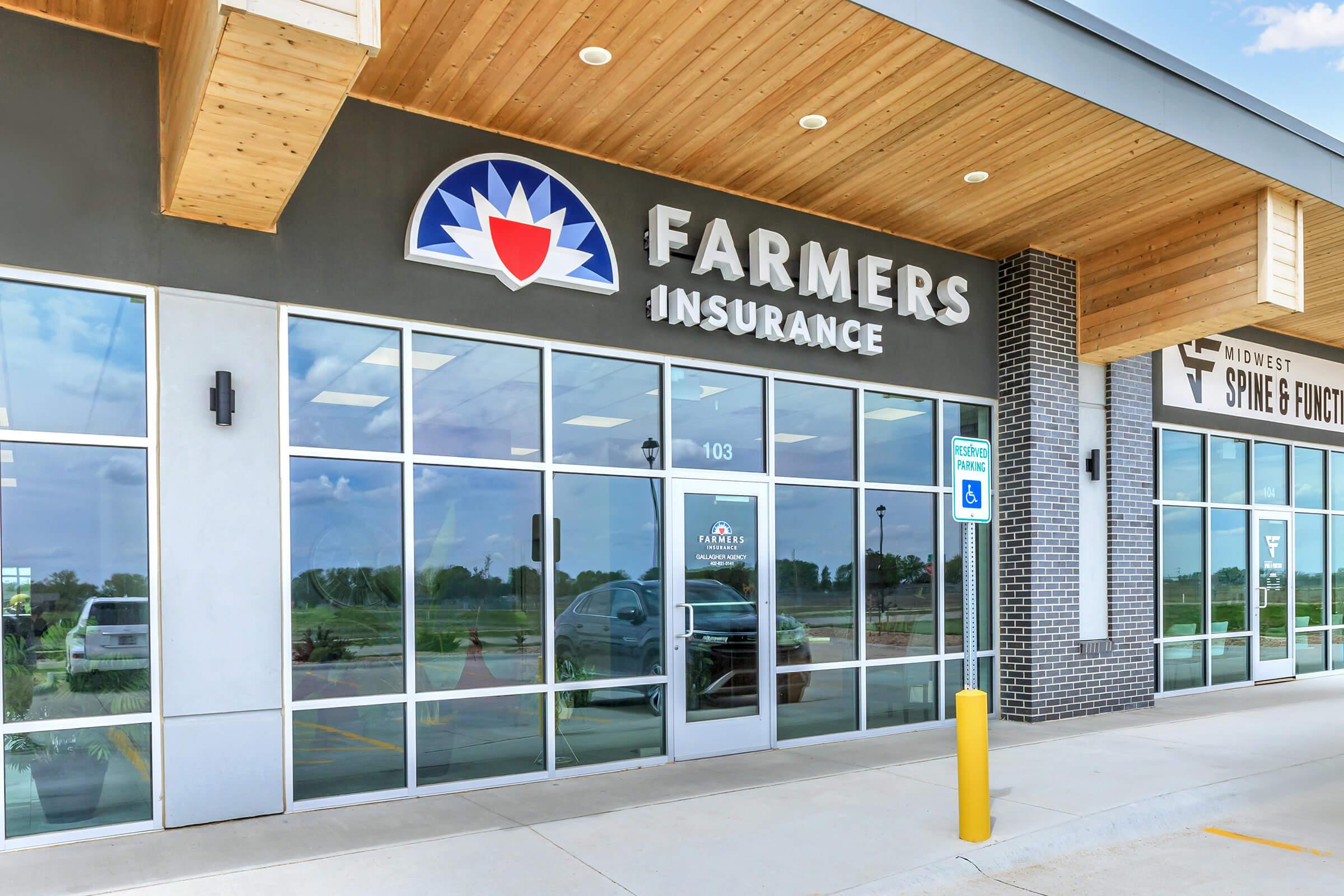 Exterior view of a Farmers Insurance office building. The entrance features large glass windows with the company’s logo prominently displayed above the door. The modern architecture includes wooden accents and a concrete pathway leading to a parking area. Blue sky visible overhead.