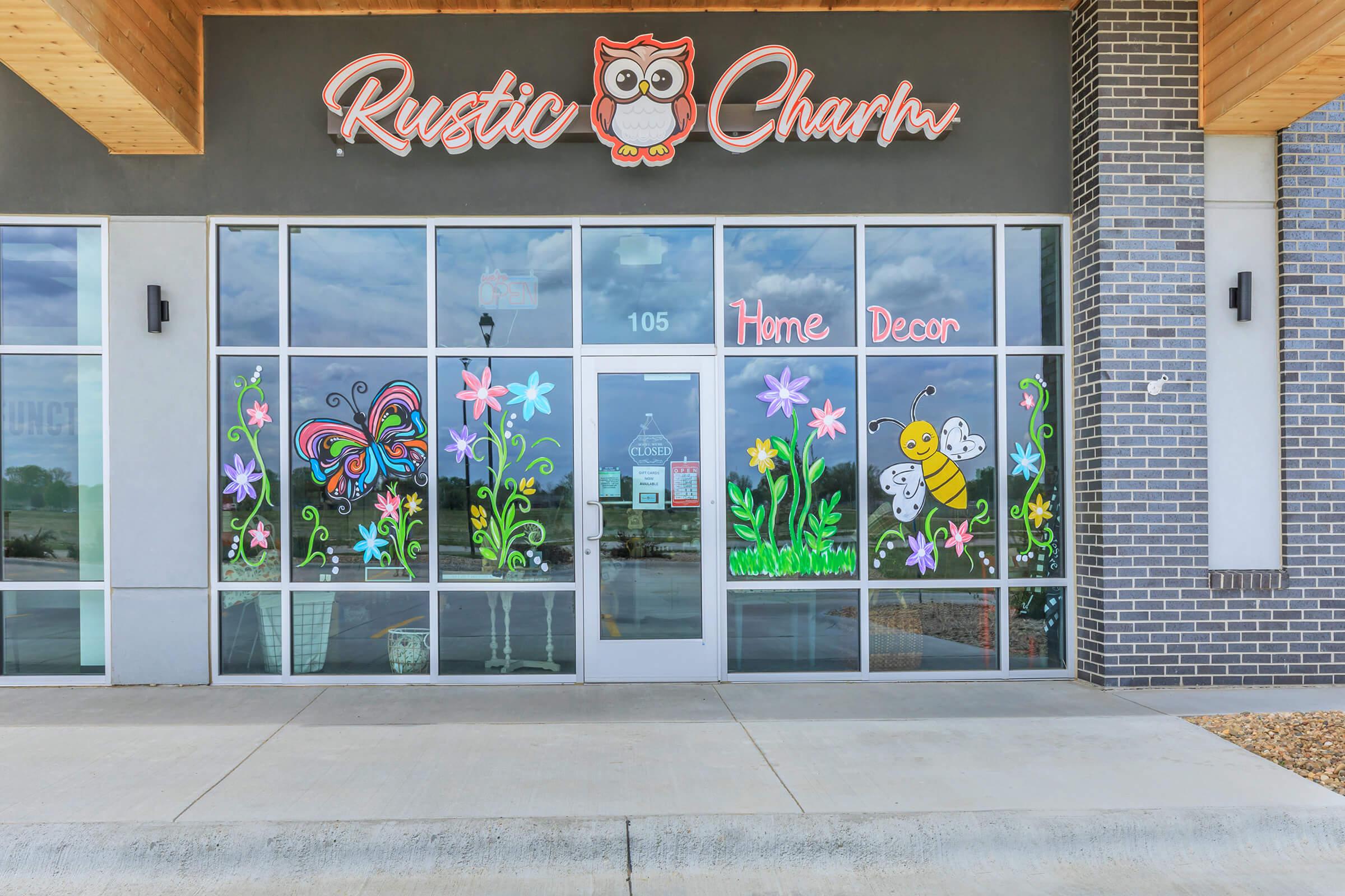 Storefront of "Rustic Charm," featuring colorful window designs of butterflies, flowers, and a bee. The shop specializes in home decor and has a welcoming vibe with artistic, hand-painted illustrations on the glass. The entrance is located in a modern building with a neutral exterior.