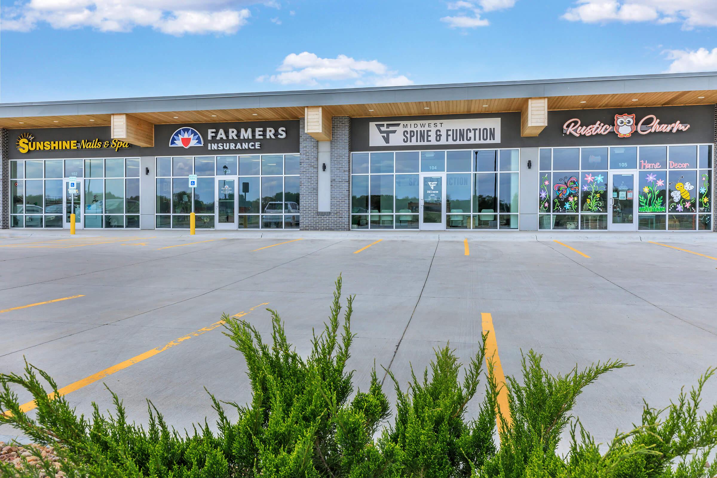 A commercial building with multiple storefronts, featuring signs for Sunshine Nails & Spa, Farmers Insurance, Midwest Spine & Function, and Rustic Charm. The parking lot is partially filled and landscaped with green bushes in the foreground, under a clear blue sky with a few clouds.