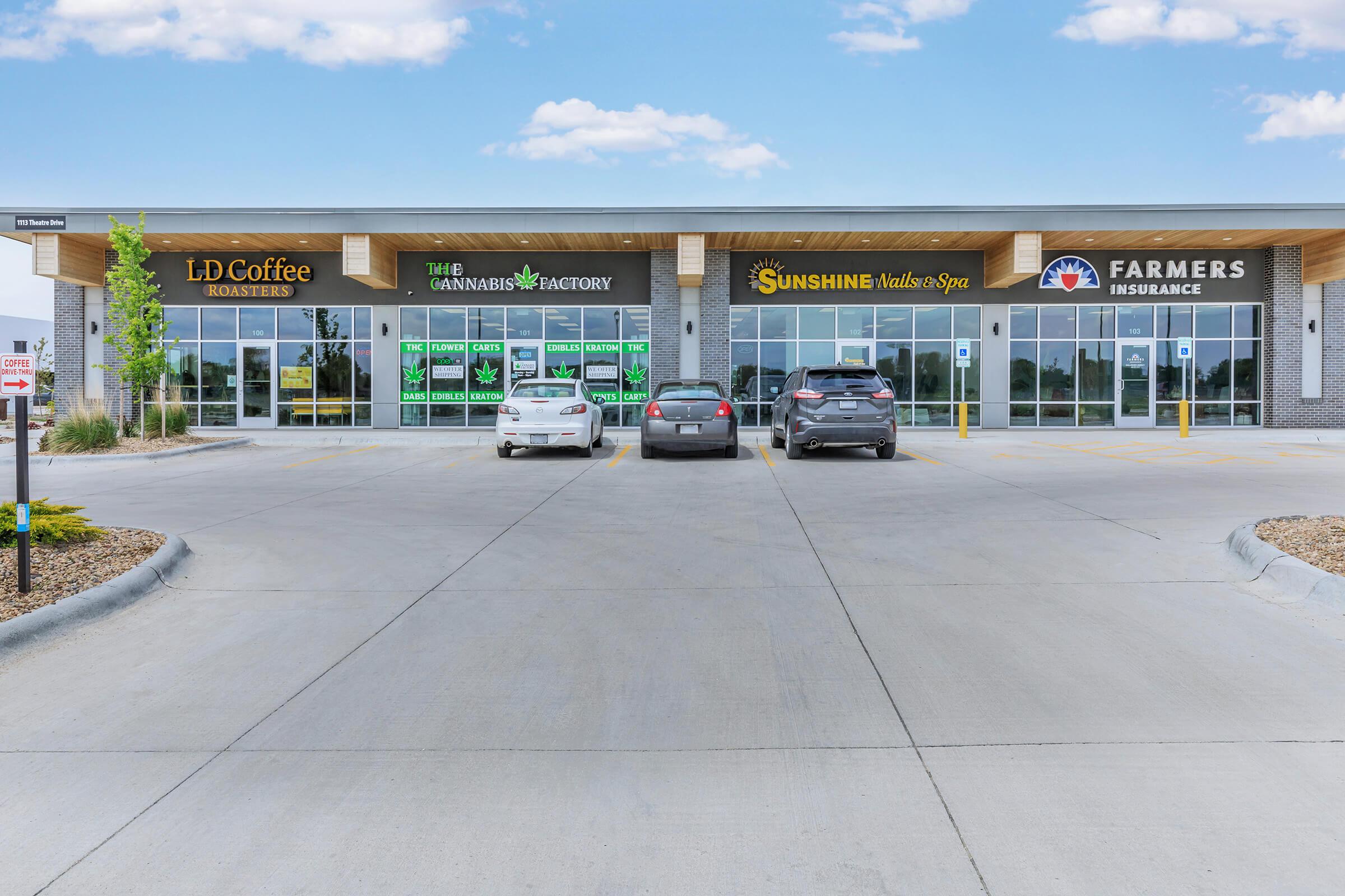 A commercial building featuring several storefronts: LD Coffee Roasters, The Cannabis Factory, Sunshine Nails & Spa, and Farmers Insurance. The parking lot is visible with parked cars, and the facade includes windows and signage for each business under a blue sky.