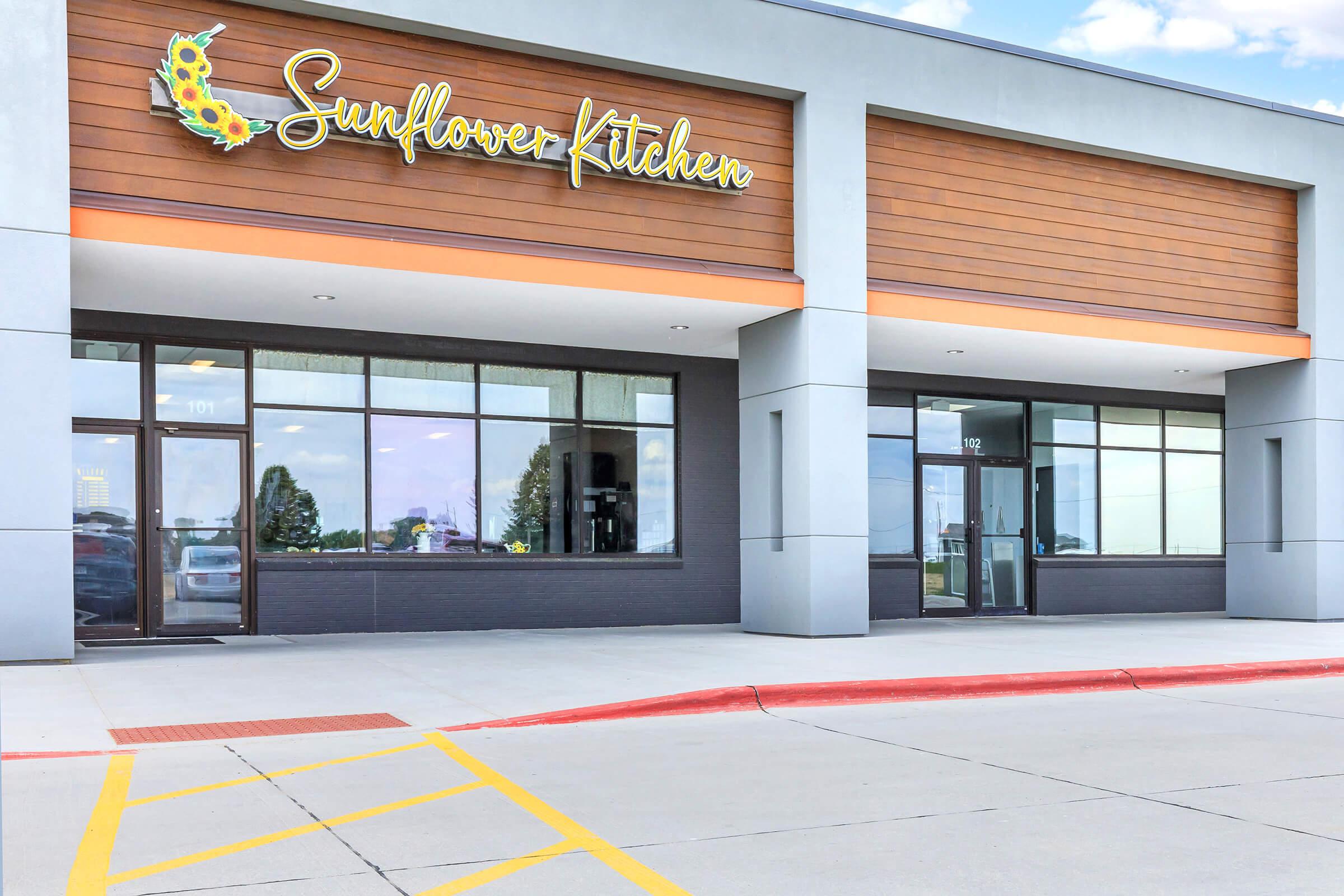 Exterior view of a restaurant called Sunflower Kitchen, featuring a modern design with large windows and a wooden facade. The sign displays the name prominently. The entrance is marked with a red curb and parking spaces visible in the foreground, under a partly cloudy sky.