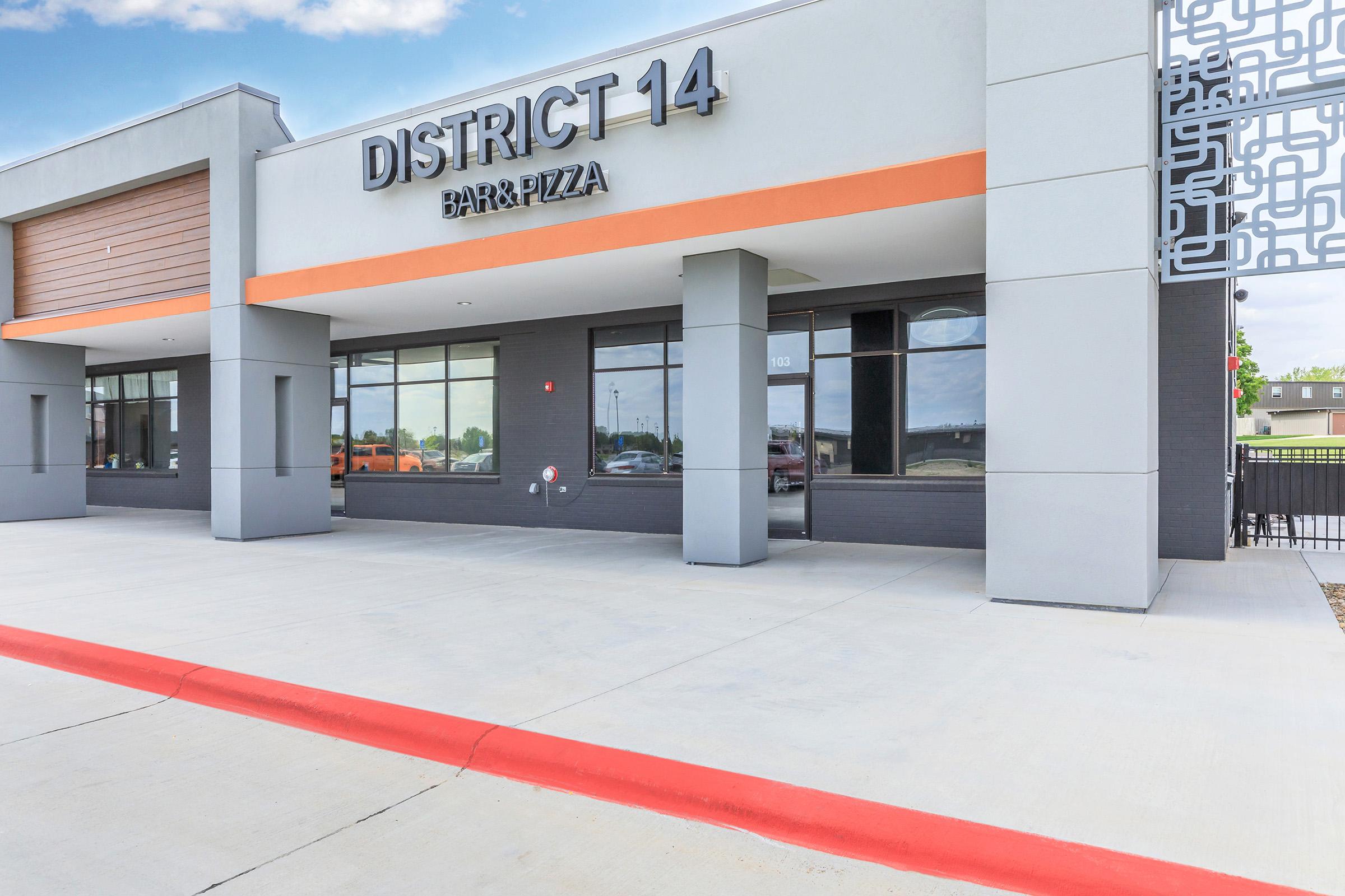 Exterior view of the District 14 Bar & Pizza building, featuring a modern design with large windows, bold signage above the entrance, and a red-curbed concrete pathway. The surrounding area is clear, with some vehicles visible in the background and green landscaping in the distance.
