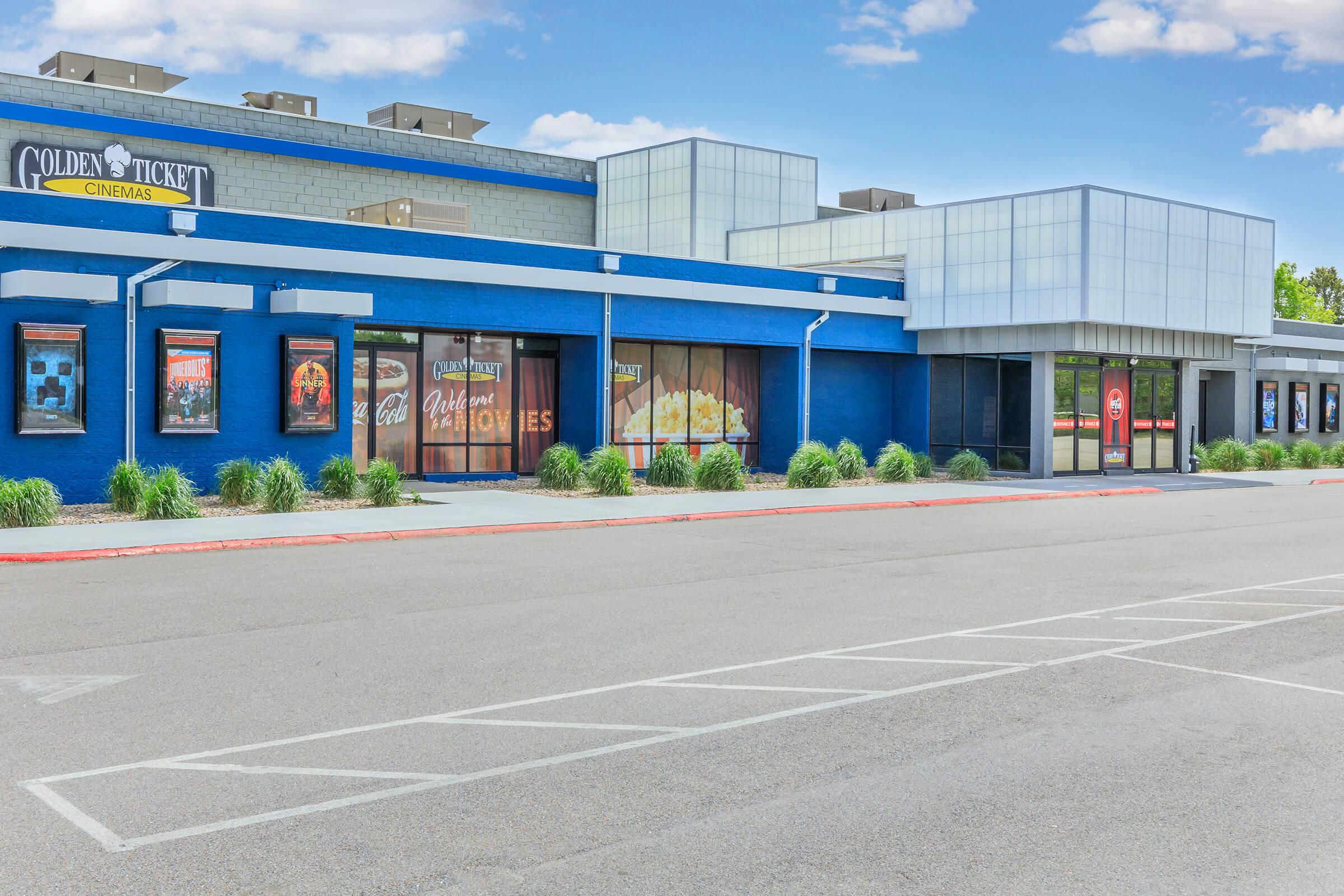 Exterior view of Golden Ticket Cinemas, featuring a blue facade, large glass windows, and promotional movie posters. The building is flanked by greenery and a clear sky. A parking lot in front showcases empty spaces, highlighting the venue's accessibility.