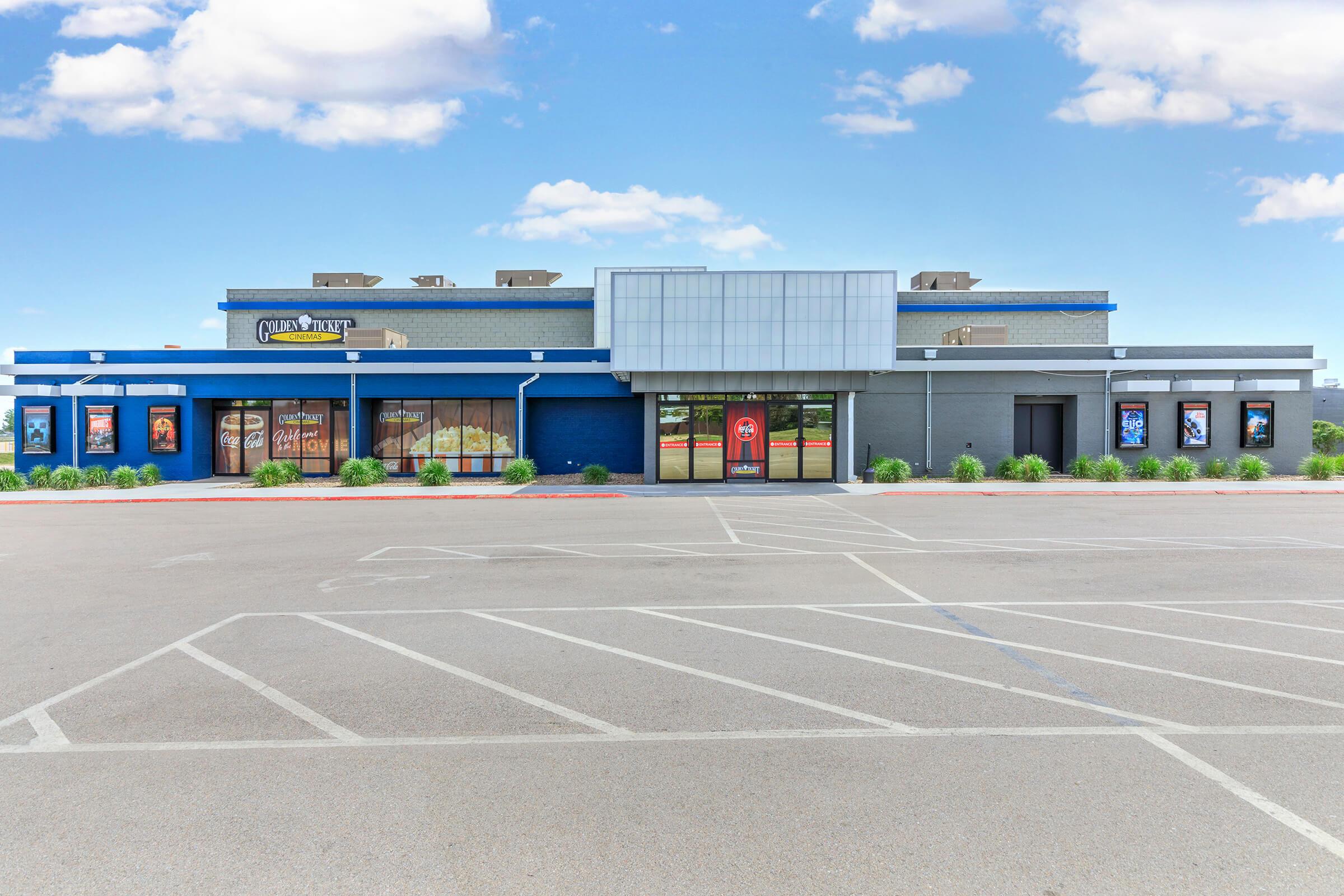 A modern building facade with a blue exterior, featuring large windows and posters advertising movies. The entrance is at the center, flanked by greenery, with a spacious parking lot in front. The sky is clear with a few clouds.
