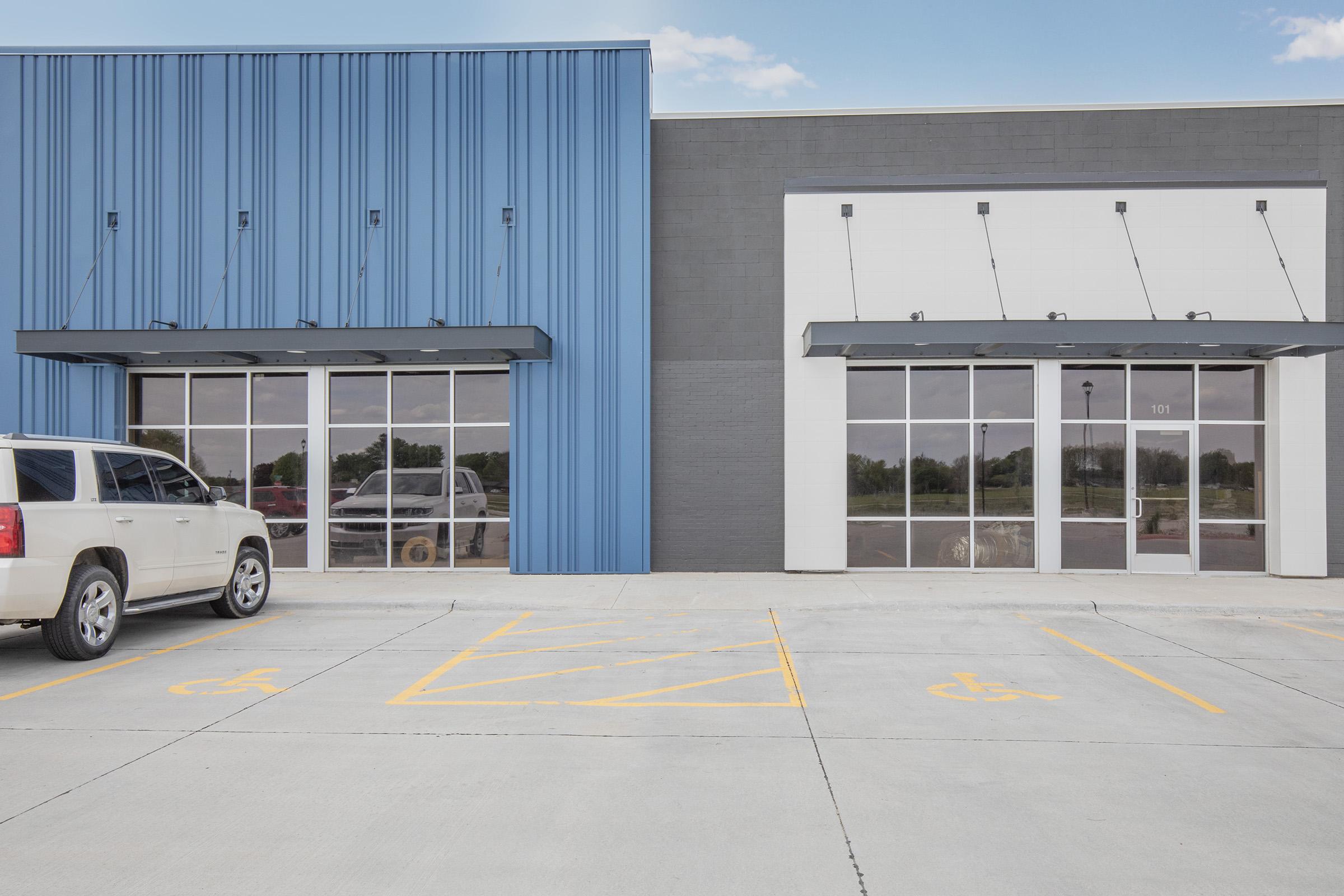 A modern commercial building featuring two storefronts with large glass windows and a sleek design. The left side is painted blue, while the right side is white and gray. In front, there is a paved parking lot with designated spaces, including accessible parking spots.