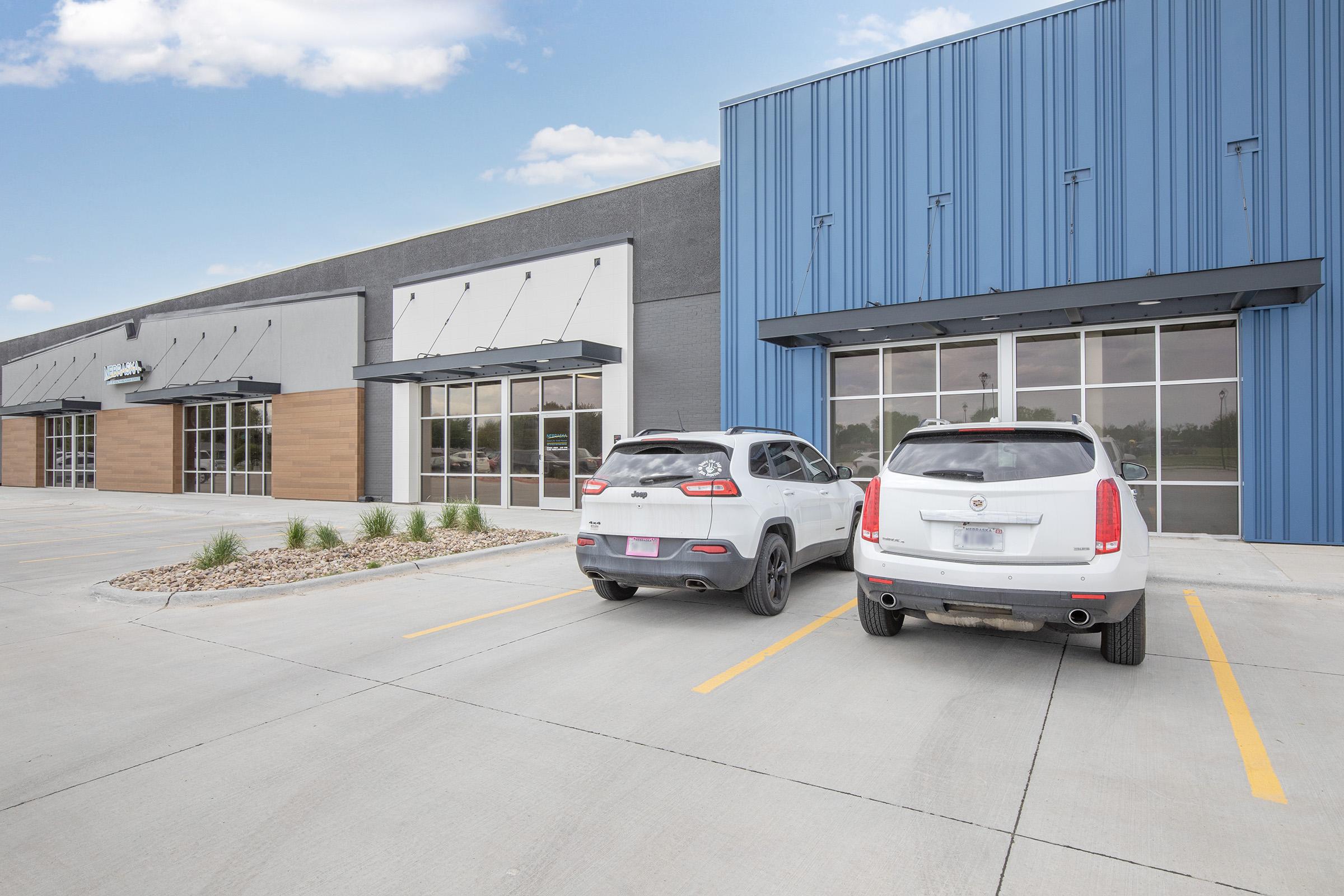 A modern commercial building with a blue exterior and large glass windows. Two parked SUVs are in the foreground, with a well-maintained asphalt parking lot featuring minimal landscaping. The sky is clear with a few clouds.