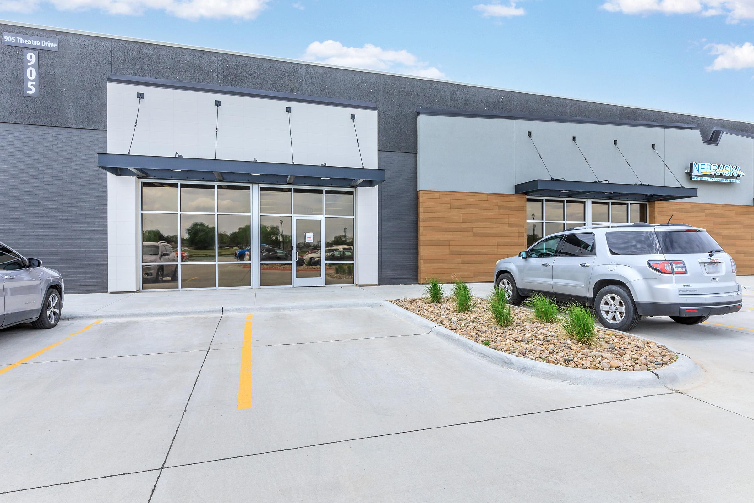 A modern commercial building exterior with large glass windows and a wooden panel entrance. The parking lot features gray and yellow lines, with two parked vehicles. Lush landscaping with rocks and grass is visible in front of the entrance, adding to the contemporary look of the property.