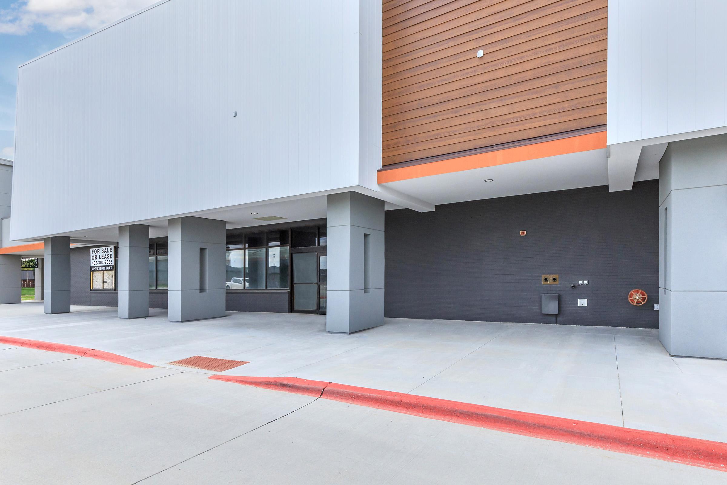 A modern commercial building exterior featuring a large, empty storefront with a gray facade and wooden paneling above. The entrance is flanked by blank windows, and the ground is paved with light-colored concrete. There are parking spaces and a red curb in front.