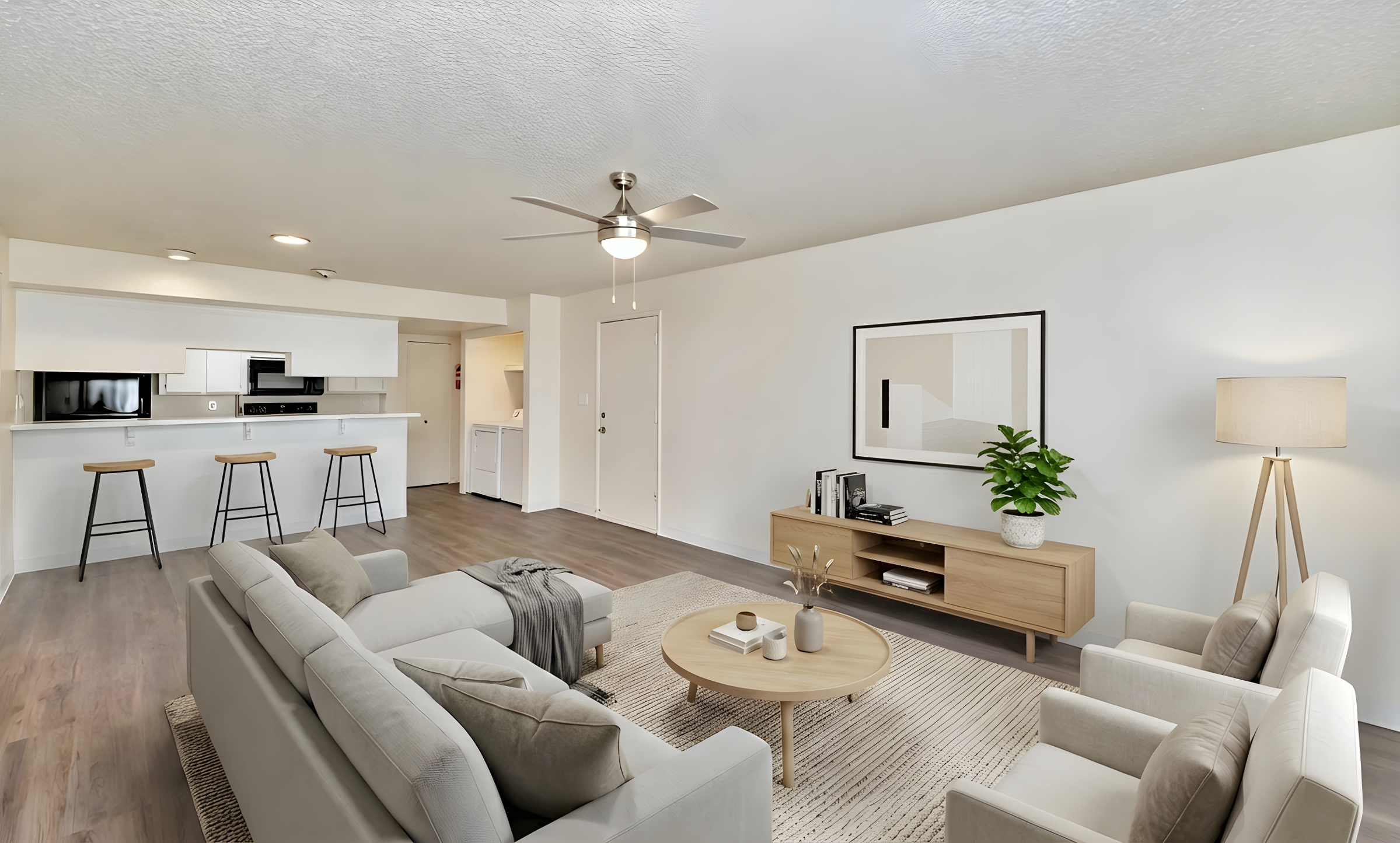 Modern living room featuring a light gray sofa, two matching armchairs, and a wooden coffee table. A large floor lamp and a potted plant add warmth. The room has light-colored walls and a ceiling fan, with a kitchenette visible in the background. Stylish decor includes a framed picture and a minimalist TV stand.