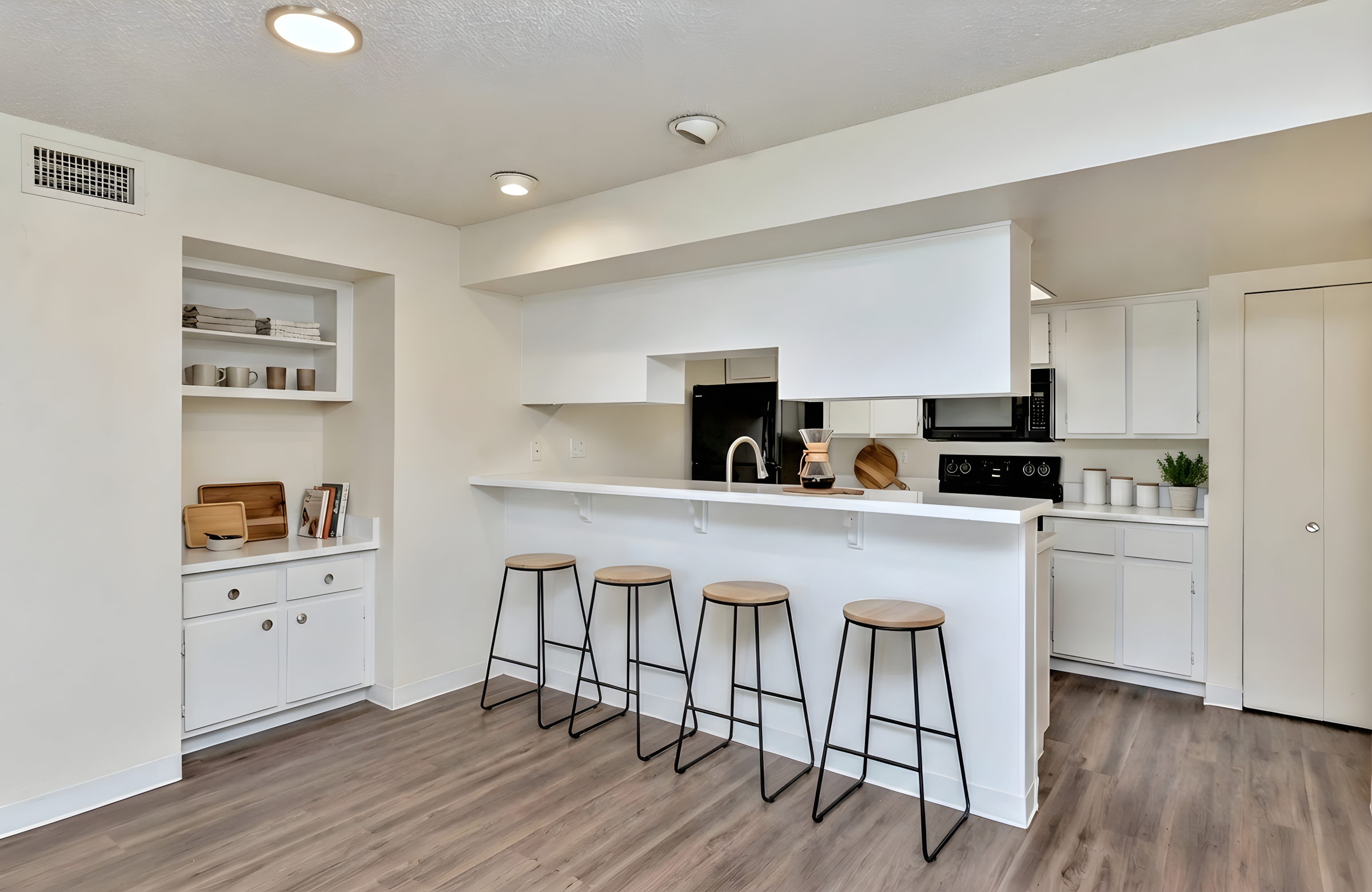 A modern kitchen featuring a white island bar with four black metal stools, white cabinetry, and hardwood flooring. Shelves with dishware are visible along the wall, and there are appliances like a black microwave and stove in the background, creating a clean and inviting space.