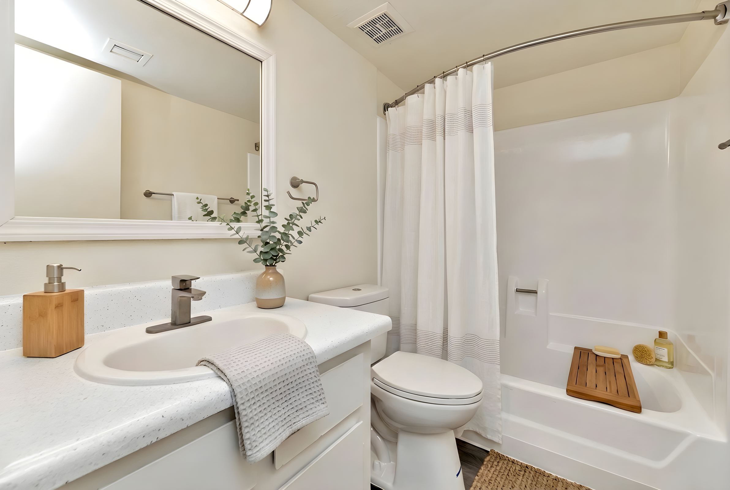 A clean and modern bathroom featuring a white sink with a faucet, a wooden soap dispenser, a small plant in a vase, a shower with a white curtain, a toilet, and a bathtub with a wooden bath tray. The walls are light-colored, and there is a mirror above the sink. Natural light filters in from a nearby window.