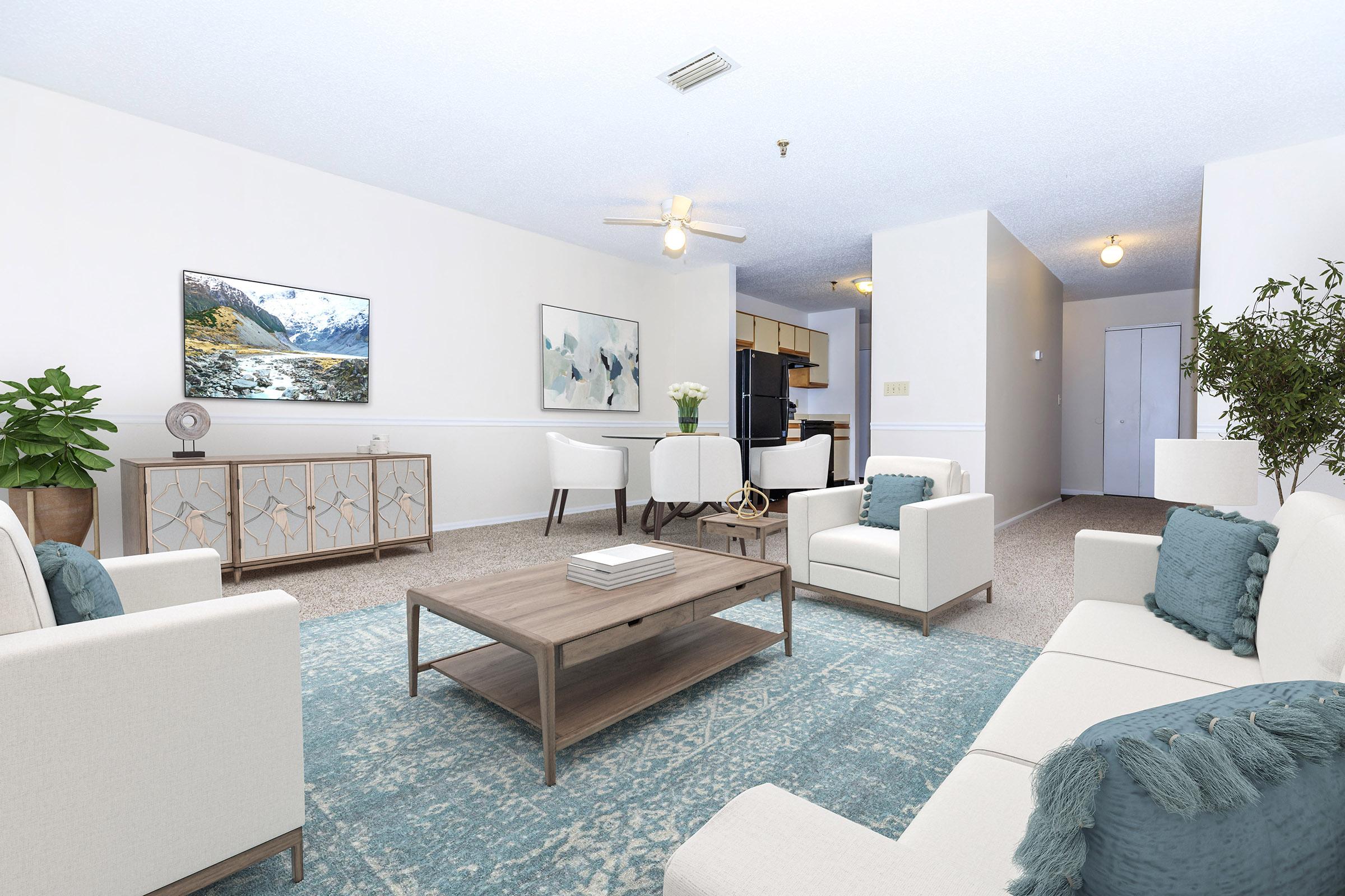 A modern living room featuring a light-colored sofa and armchairs, a wooden coffee table, and a large area rug with a blue pattern. The space includes a flat-screen TV mounted on the wall, a plant, and a dining area with white chairs. The walls are painted in a soft color, creating a bright and airy atmosphere.