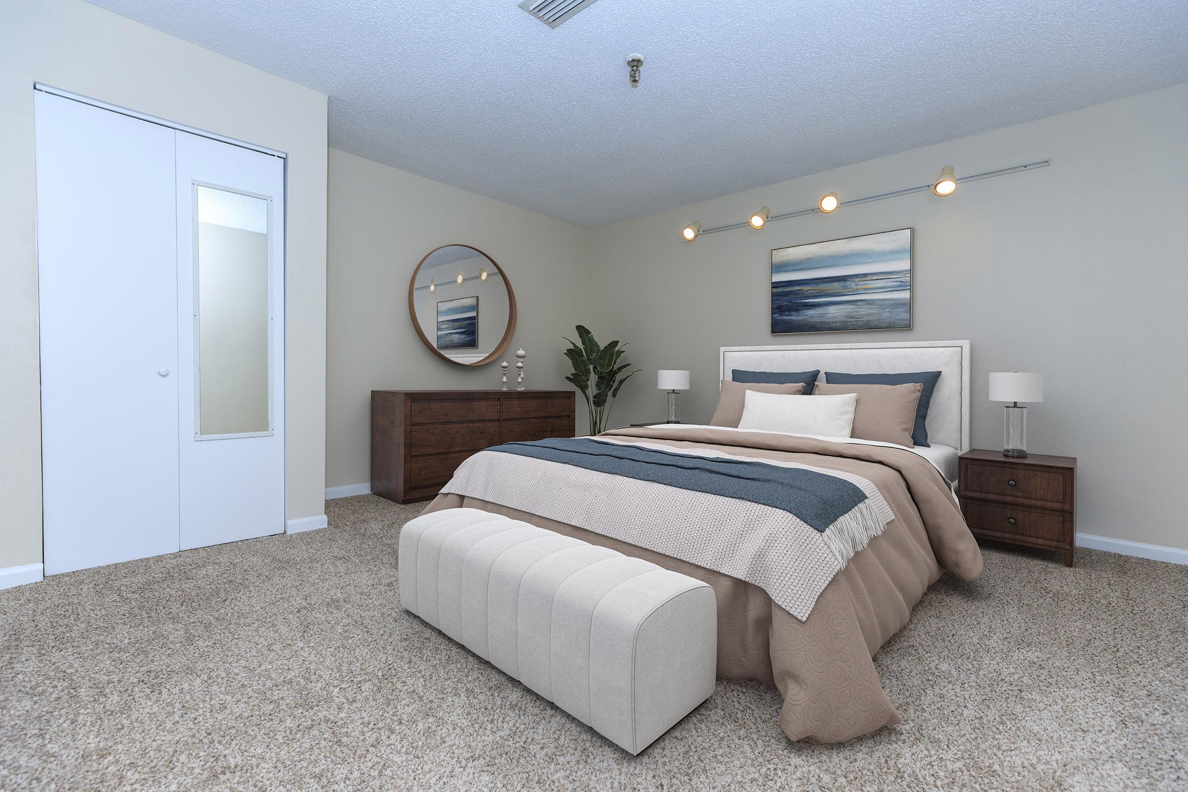 A cozy bedroom featuring a queen-sized bed with neutral bedding and decorative throw pillows. There's a round mirror above the headboard and two bedside tables with lamps. A wooden dresser is against one wall, and a bench at the foot of the bed provides extra seating. Indoor plants add a touch of greenery.