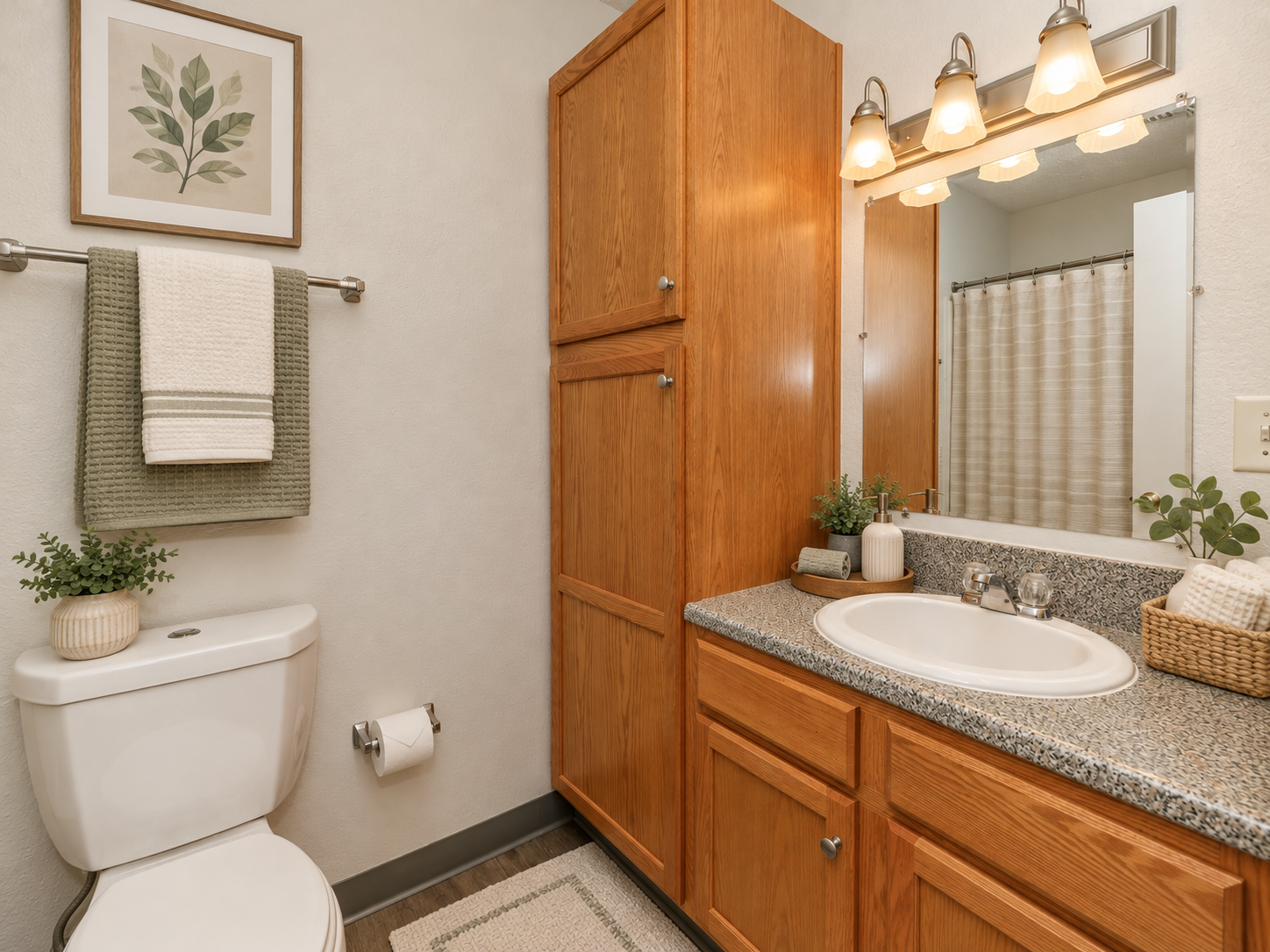 A neatly arranged bathroom featuring a white toilet, wooden cabinetry, a sink with a granite countertop, and a mirror above the sink. Decor elements include a framed leaf print, neatly hung towels, and small plants. The shower curtain is a light, textured fabric, contributing to a clean and inviting atmosphere.