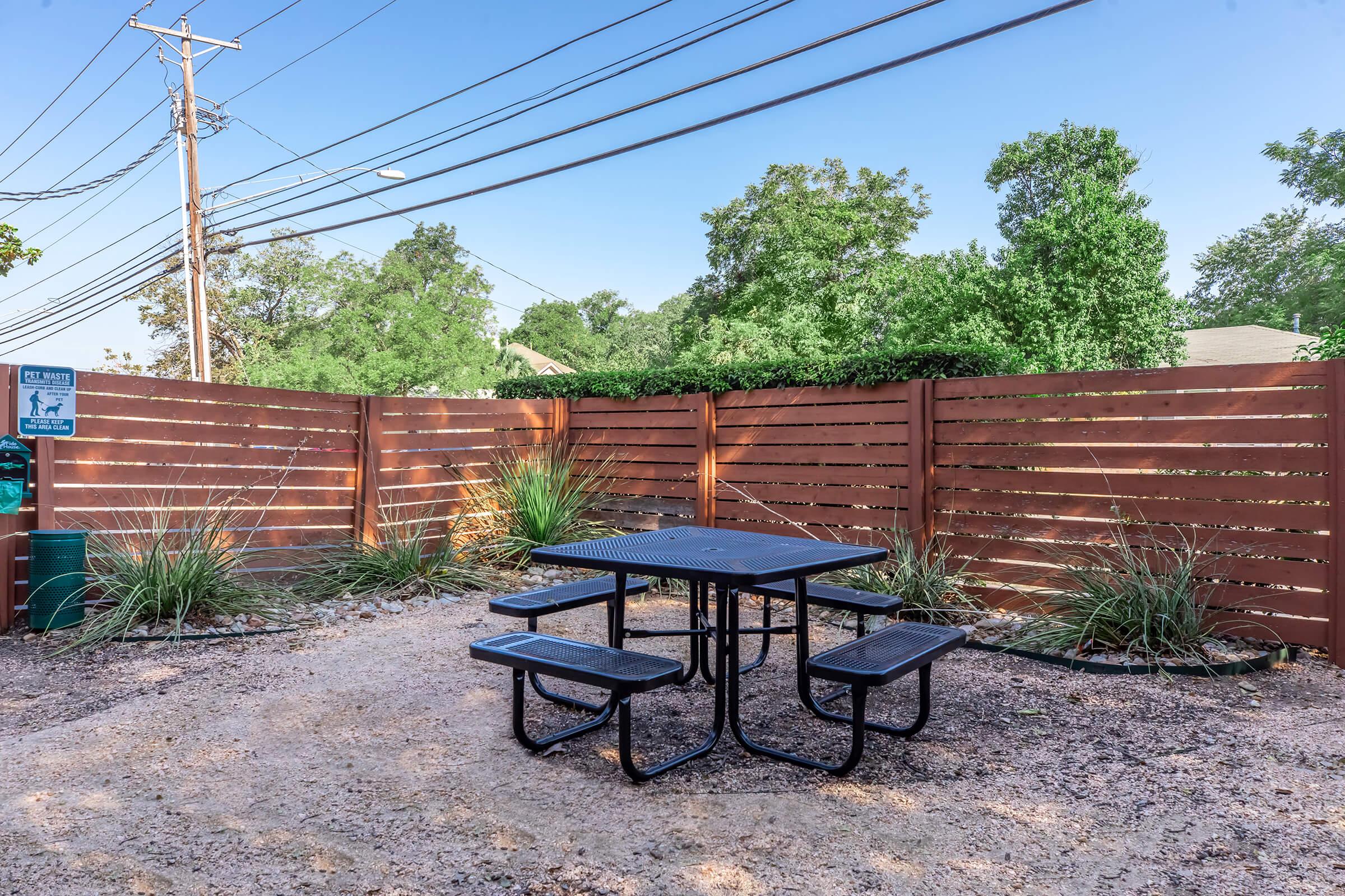 A black metal picnic table with attached benches is set in a gravel area surrounded by greenery and wooden fences. Power lines are visible overhead, and there is a sign indicating pet rules nearby. The setting is bright and sunny, creating a welcoming outdoor space.