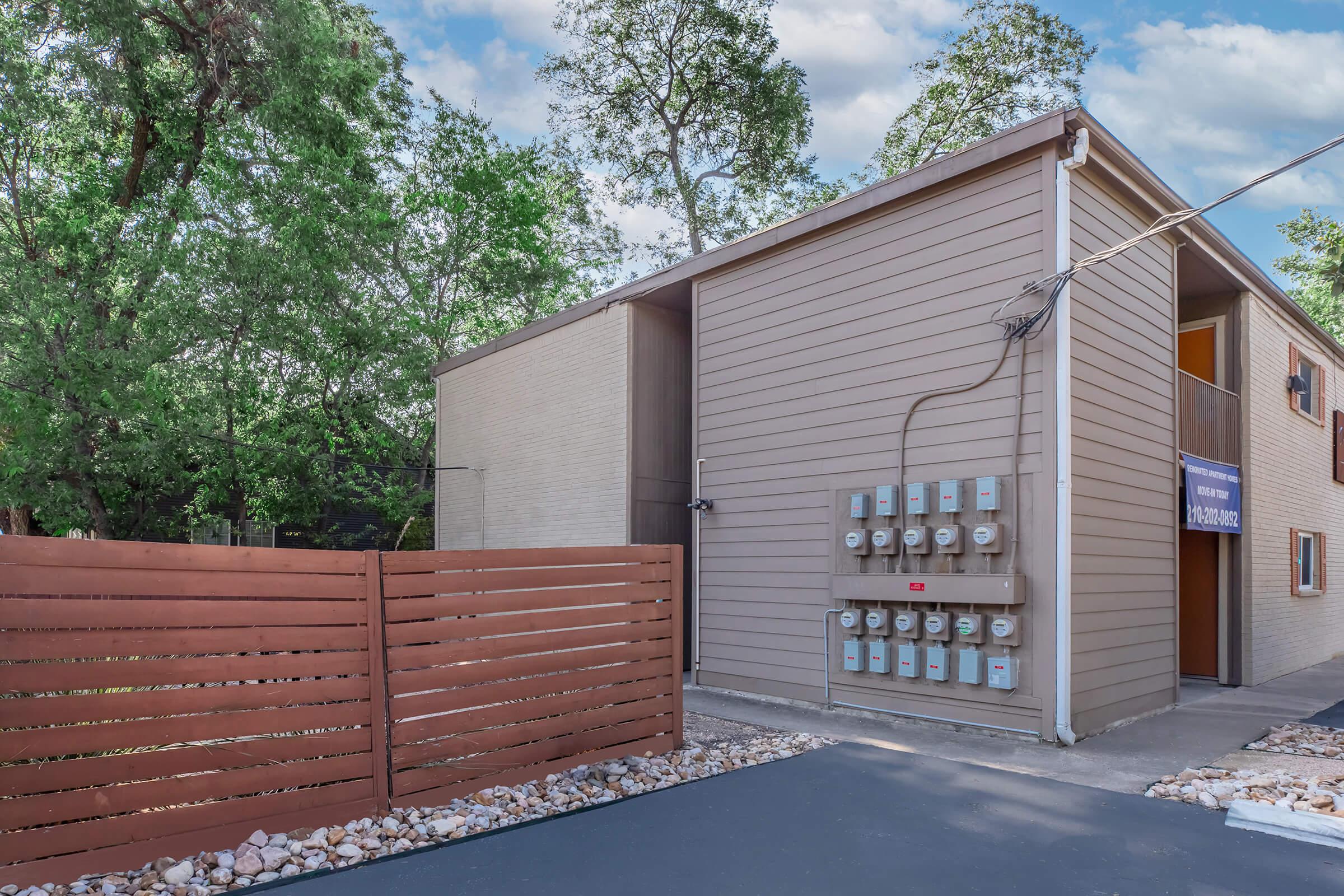 A modern building with light-colored siding, featuring multiple utility meters on the exterior wall. In the foreground, there is a wooden fence with a sleek design. Lush green trees are visible in the background, providing a natural setting. The sky is partly cloudy, adding to the inviting atmosphere.