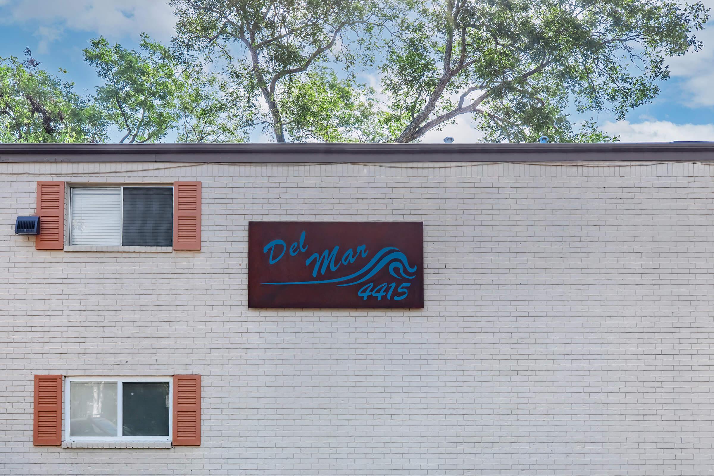 Sign on a building displaying the text "Del Mar 4415" in a stylized font, accompanied by a wave graphic. The building has light-colored brick walls and two windows with orange shutters. Green trees and a blue sky are visible in the background.
