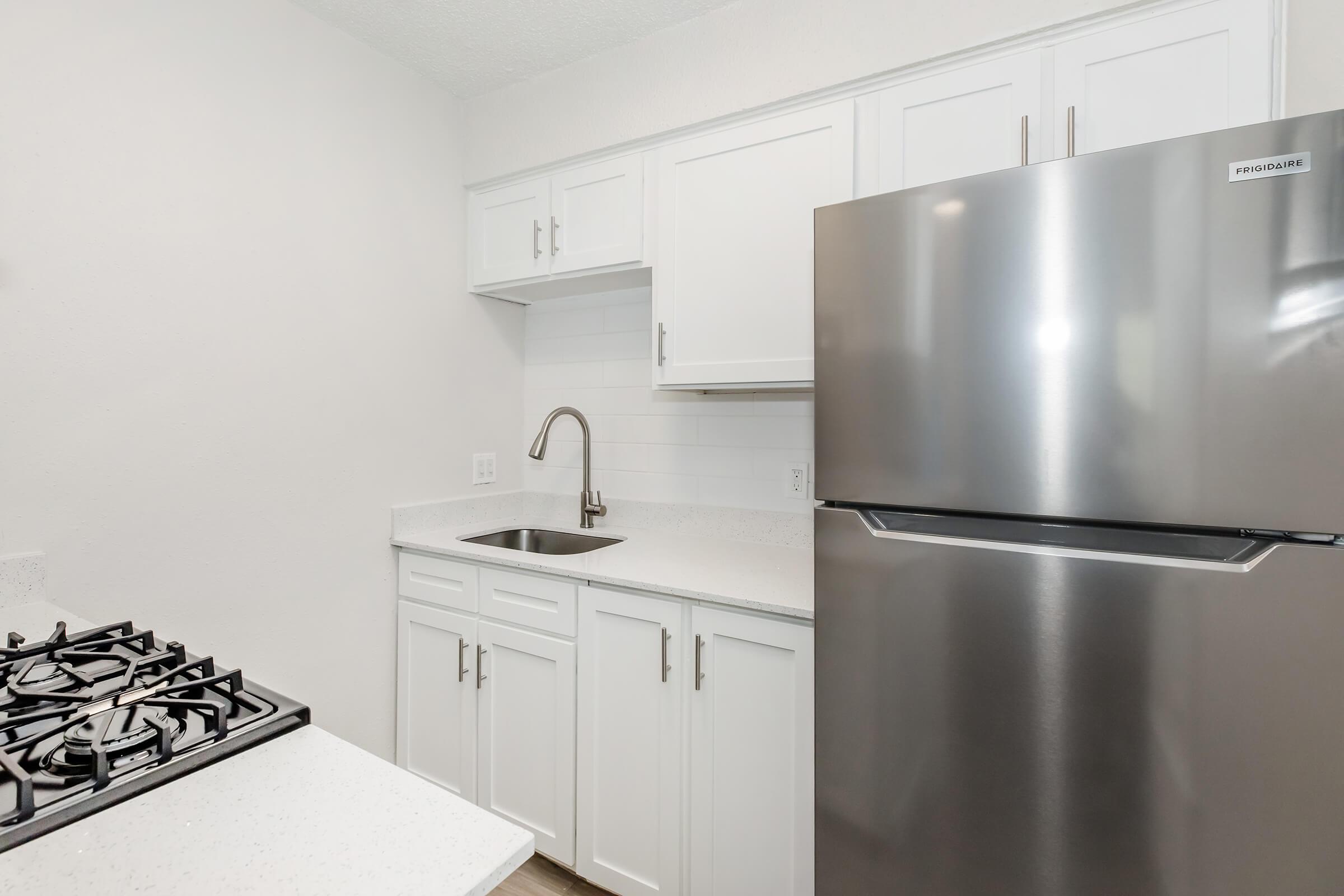 Modern kitchen featuring stainless steel refrigerator and gas stove, white cabinets, and a small sink. The countertop is light-colored and the walls are painted in a neutral tone, creating a bright and clean atmosphere.