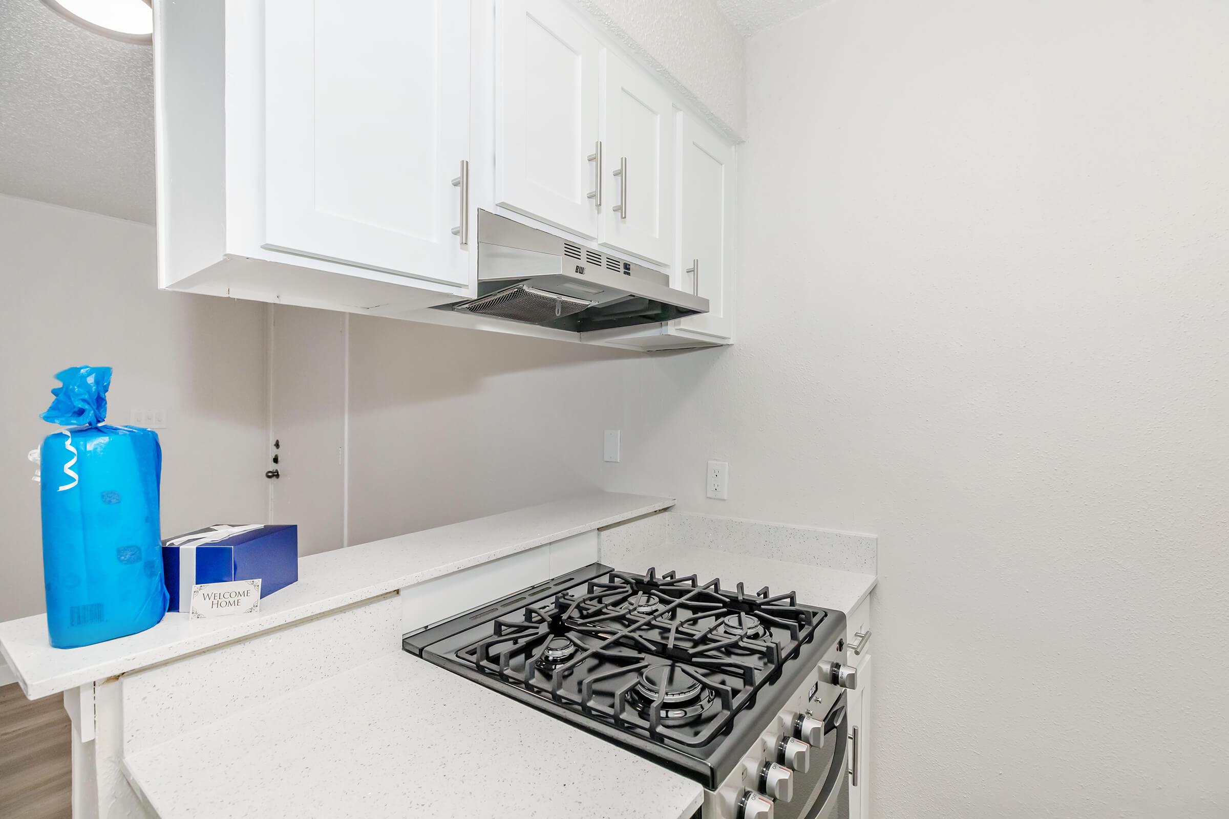 A modern kitchen featuring white cabinetry, a countertop with a gas stove, and an overhead range hood. To the left, there is a blue plastic bag and a white box on the counter. The walls are painted a light color, enhancing the bright and clean appearance of the space.