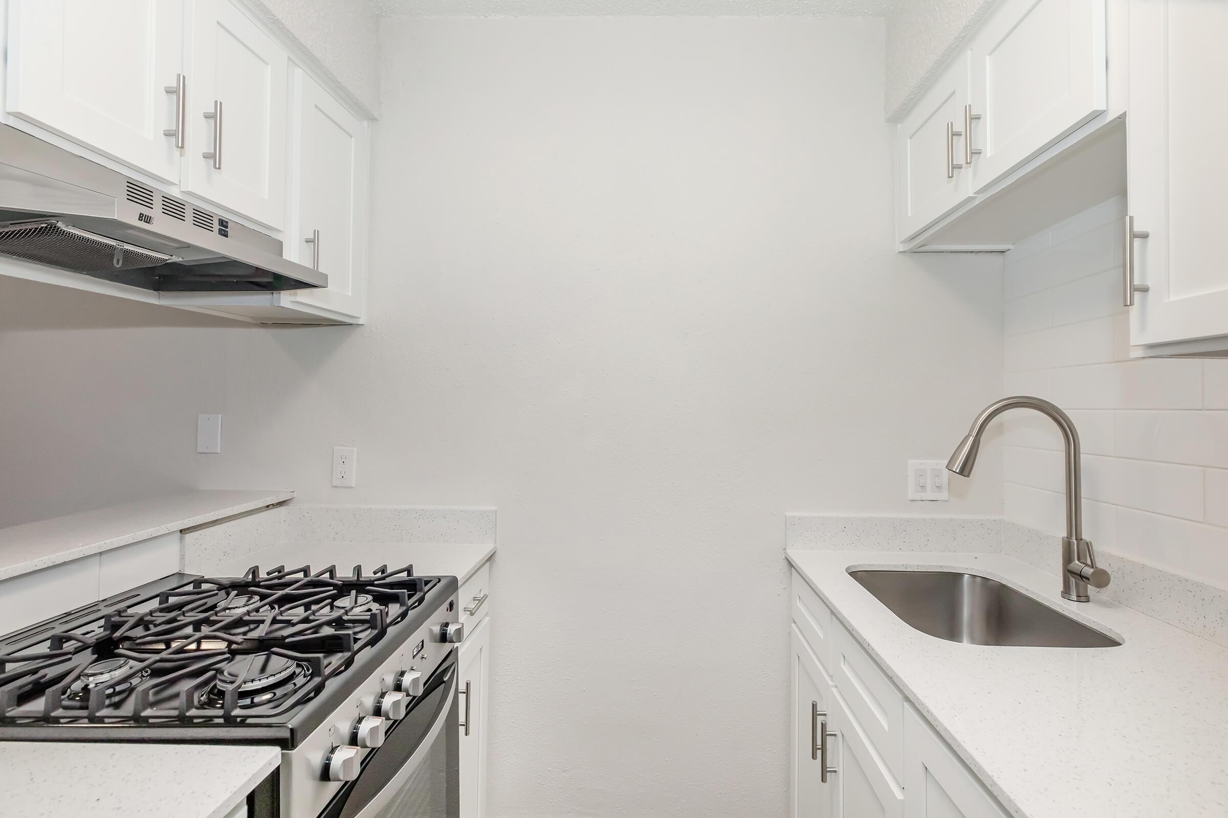 A modern kitchen featuring white cabinetry, a stainless steel gas stove, and a sleek sink. The walls are light-colored, and there is a white tiled backsplash. The space is well-lit, creating a clean and bright atmosphere.