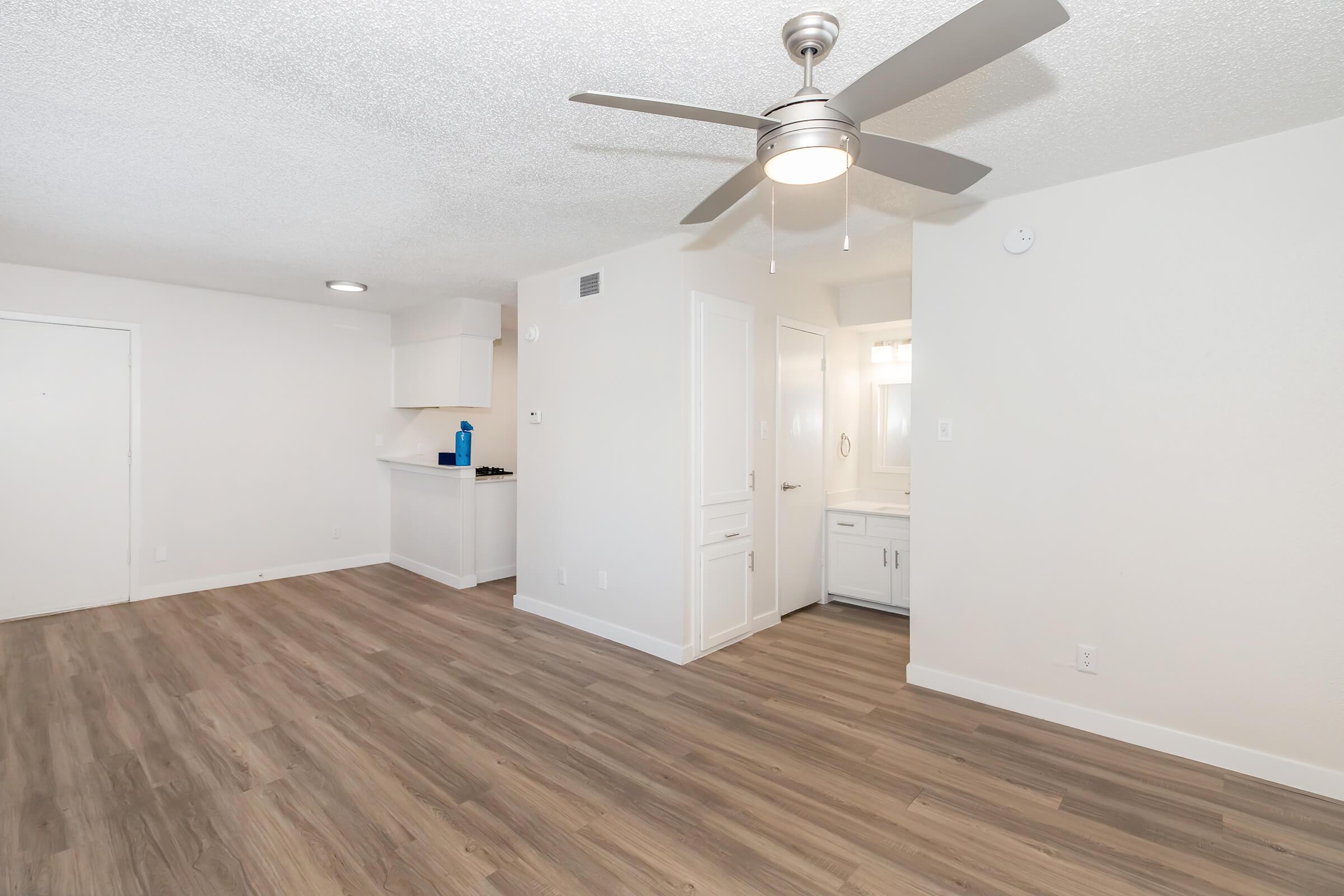 Spacious, modern living area featuring light-colored walls, wood-style flooring, and a ceiling fan. In the background, there is a kitchenette with a white countertop and entrance to a bathroom area. The space is well-lit with natural light and has a clean, minimalistic design.