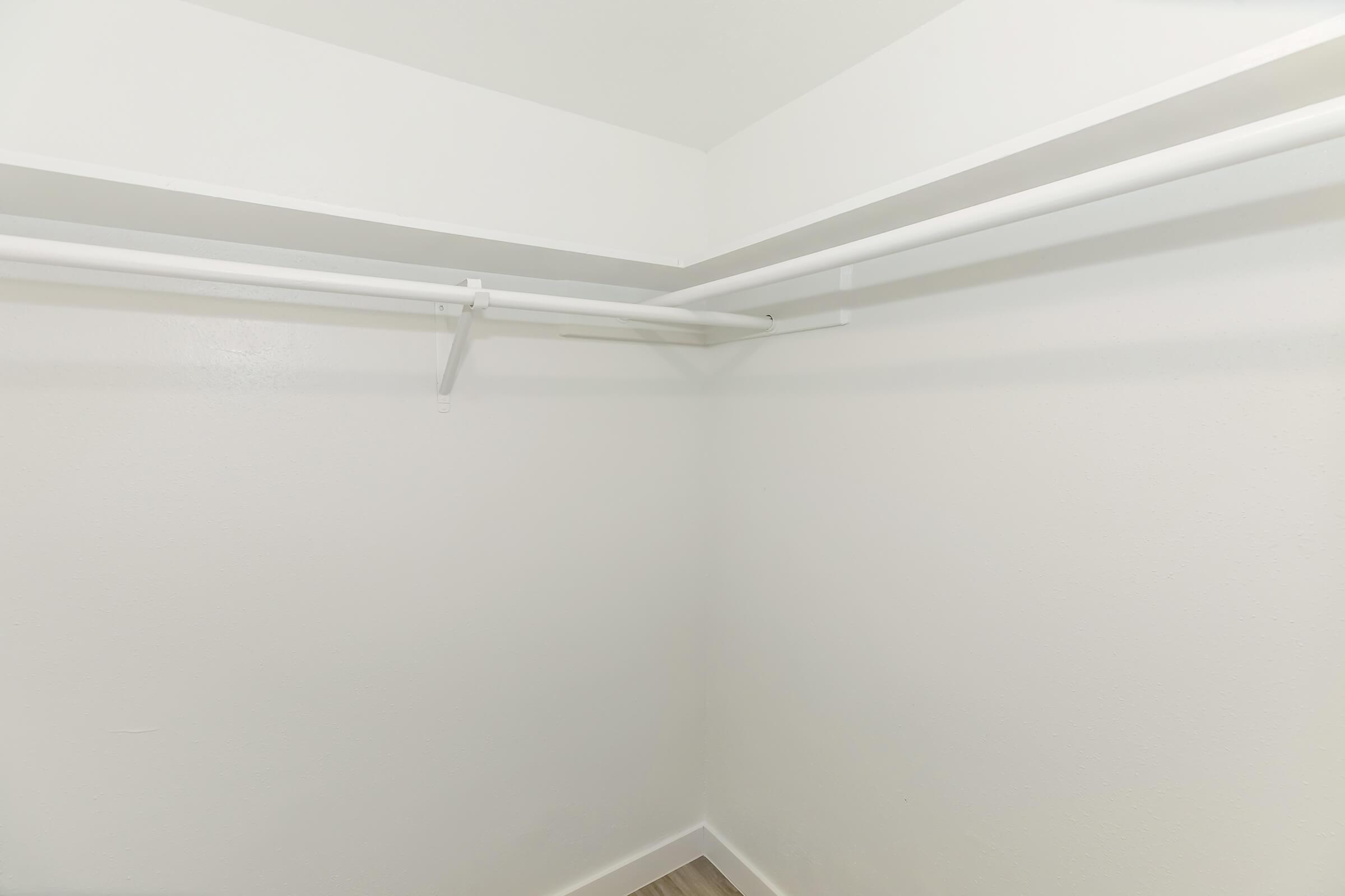A minimalist closet with white walls and two horizontal white rods for hanging clothes. The floor is wooden, and there is ample space for storage, but the closet is currently empty.