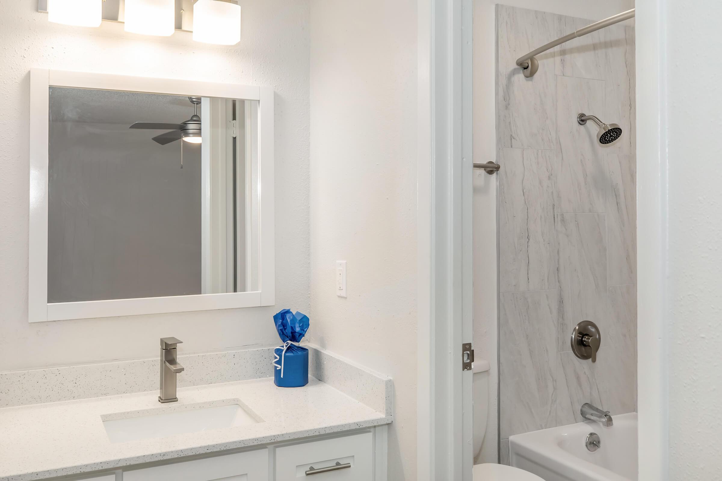 A modern bathroom featuring a white countertop sink with a sleek faucet, a large wall mirror, and a shower area with marble tiles. A blue decorative item sits on the counter, and a light fixture above provides illumination. The space is clean and minimalistic with neutral colors.