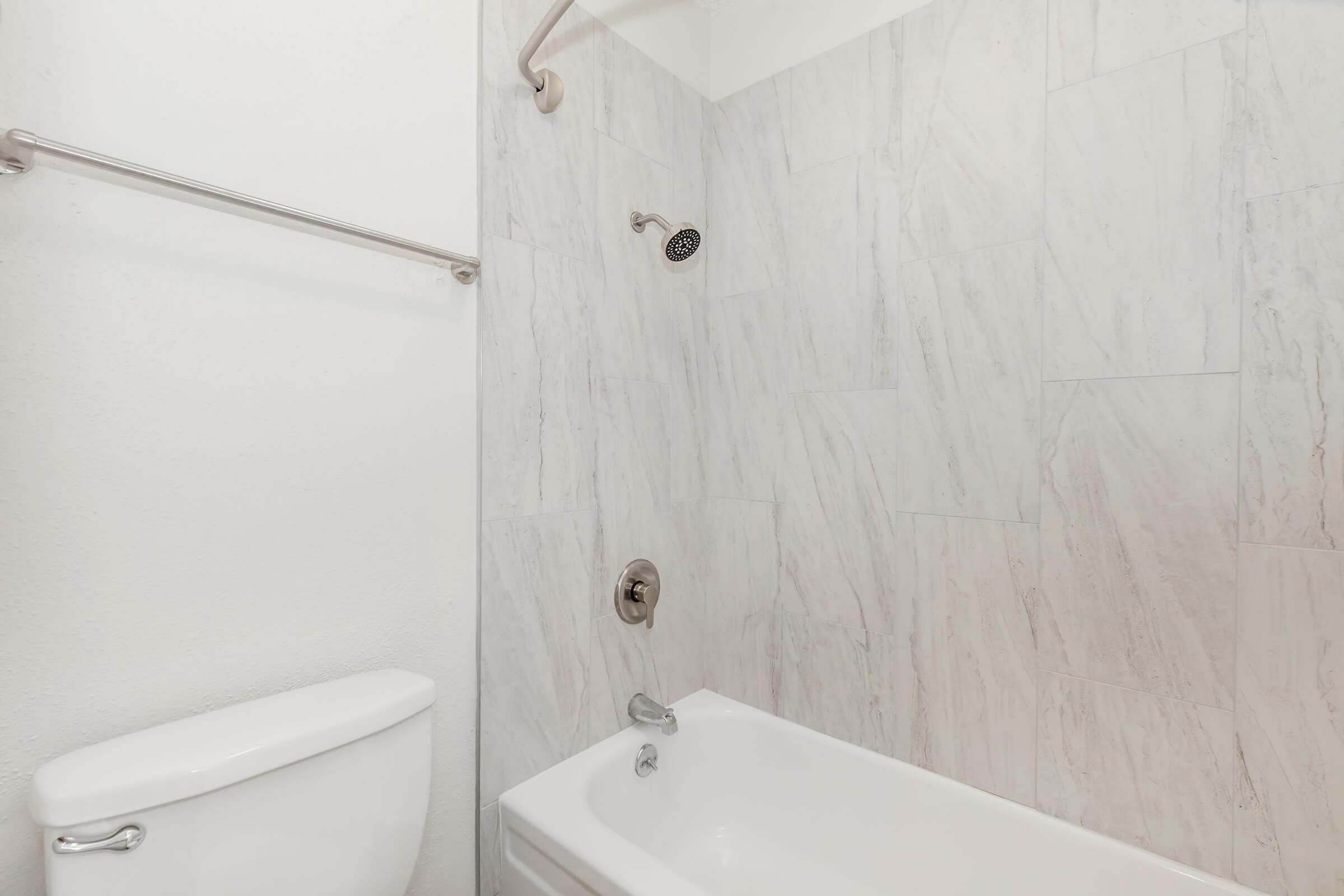 A clean and minimalistic bathroom featuring a white toilet, a bathtub with a showerhead above, and light-colored marble-style wall tiles. The overall design is modern and functional, emphasizing a bright and airy space.