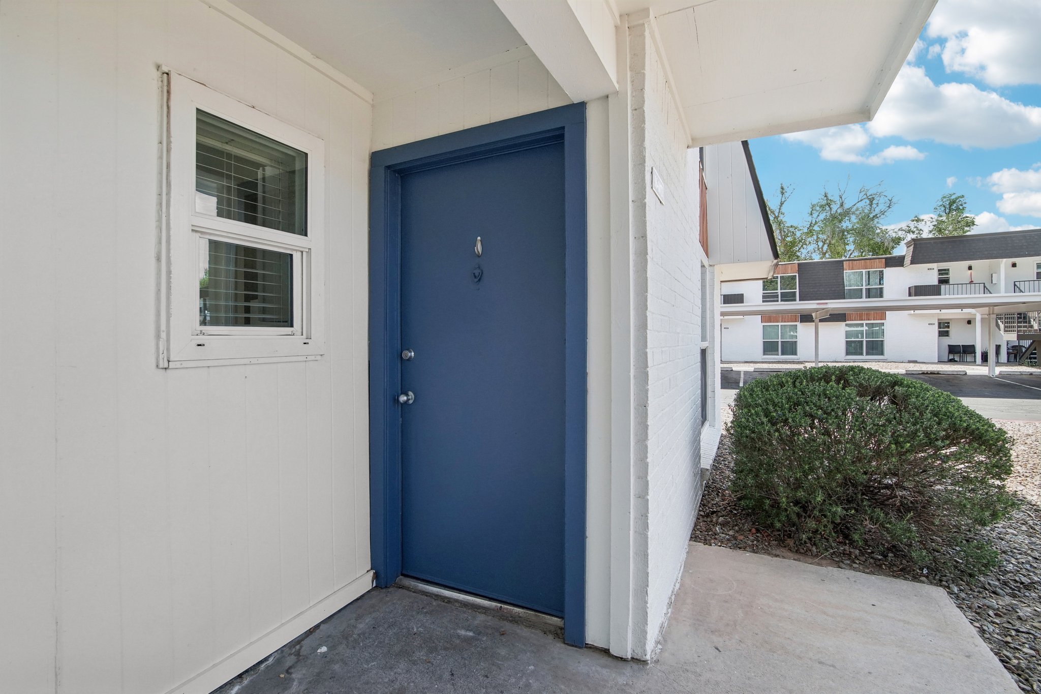 A blue front door in a white and light gray building. The door is flanked by a small window, and there is a small bush nearby. In the background, a low-rise building with multiple units can be seen, under a partly cloudy sky. The setting appears to be a residential area.