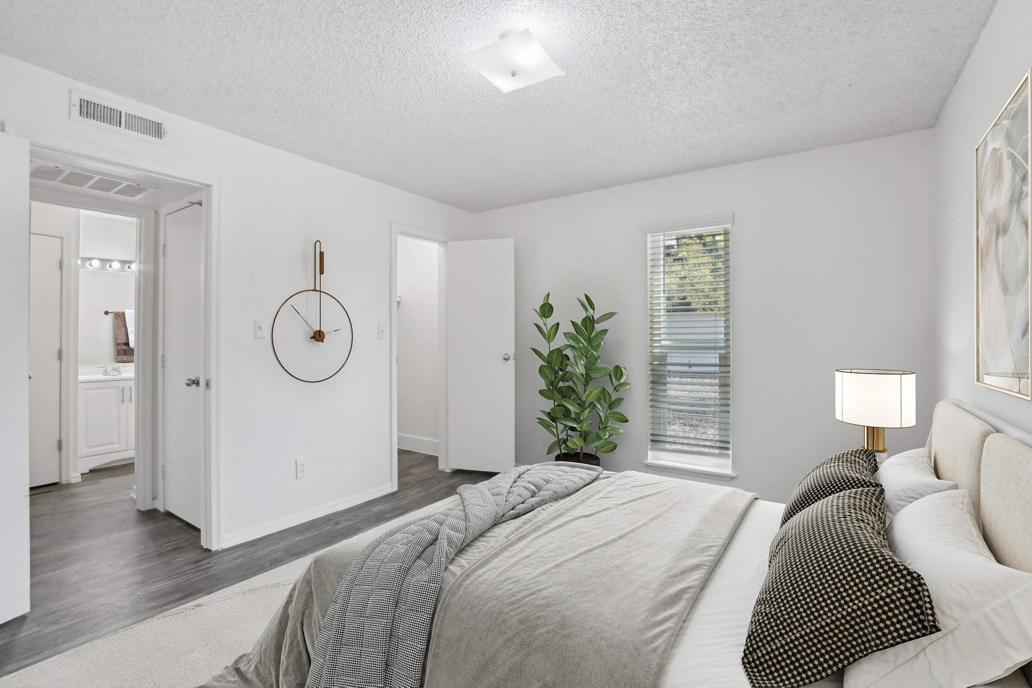 Bright and airy bedroom featuring a light-colored wall, a large bed with textured bedding and accent pillows, a stylish bedside lamp, and a modern clock on the wall. A potted plant adds a touch of greenery, and there is a doorway leading to a bathroom with a mirror visible. Natural light streams through a window.