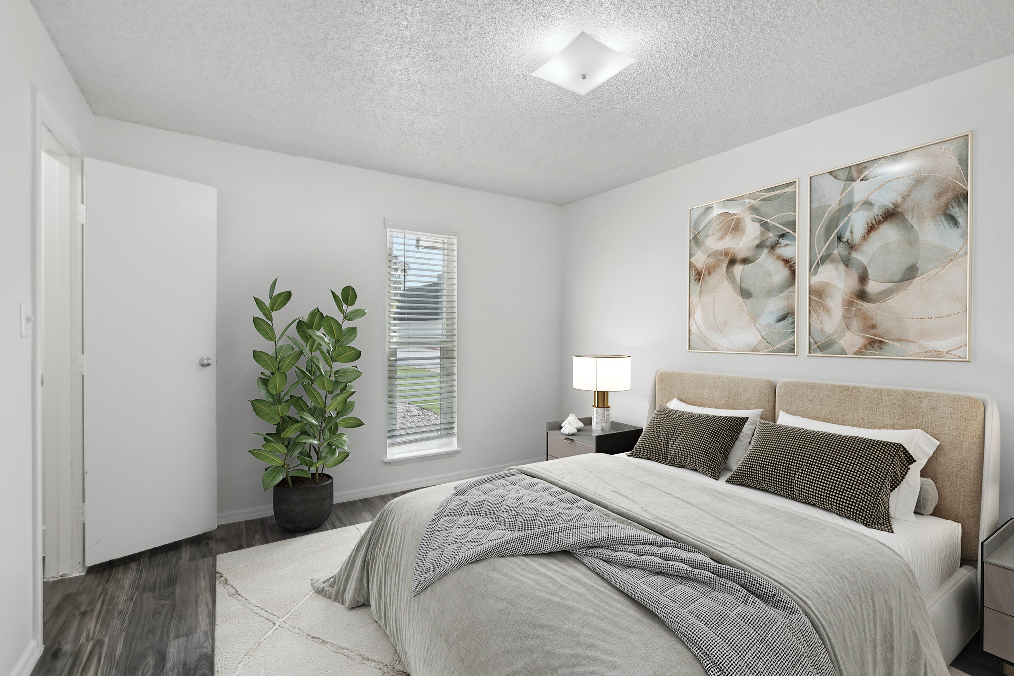 A modern bedroom featuring a neatly made bed with light bedding, two decorative pillows, and a textured throw blanket. There is a small nightstand with a lamp beside the bed, a tall green plant in the corner, and two abstract art pieces on the wall. Natural light comes through a window, and a door leads to another room.
