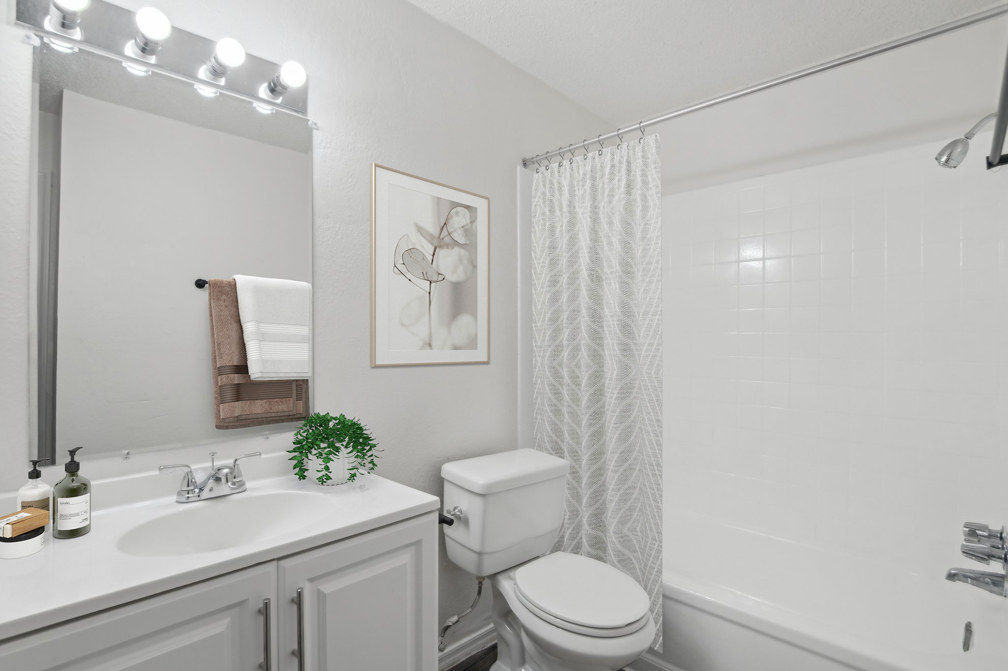 A modern bathroom featuring a white bathtub with a shower curtain, a white toilet, a vanity with a sink and mirror, and decorative elements like a framed artwork and a small plant. Soft lighting from a mirror-mounted fixture enhances the contemporary aesthetic.