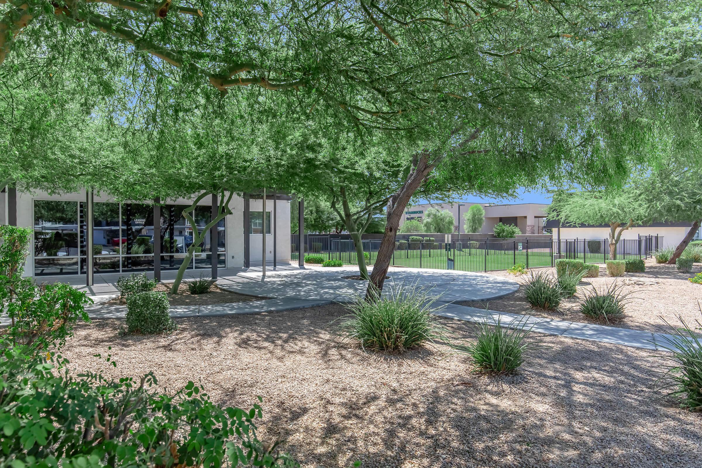 Modern building surrounded by lush greenery and trees, featuring a paved pathway and landscaped areas with gravel. In the background, there are fences and additional greenery, creating a tranquil outdoor environment. Bright blue sky visible above the trees.
