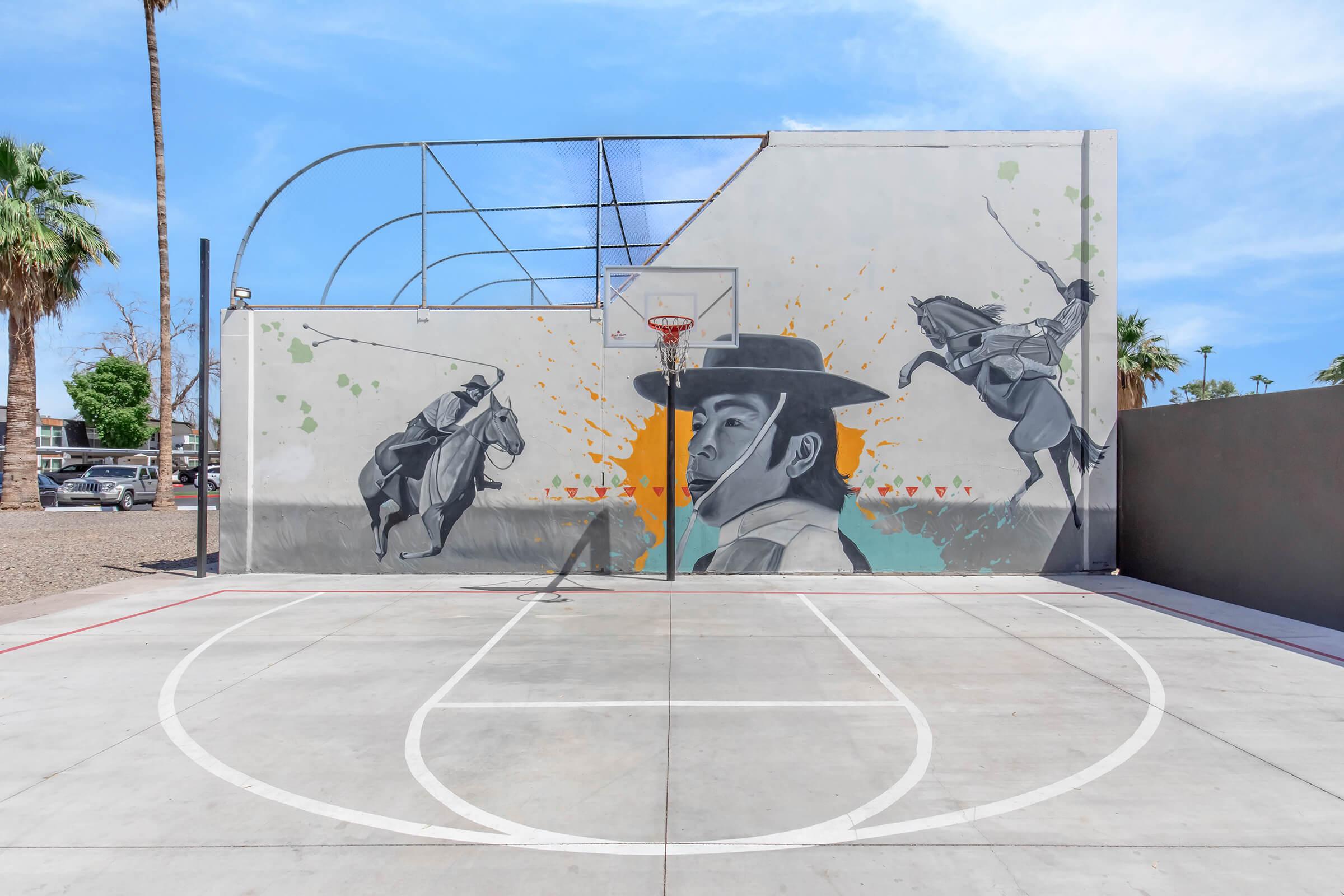 A basketball court with a colorful mural on the wall depicting a man wearing a hat alongside two horse riders. The mural features vibrant colors and artistic details, surrounded by palm trees and a clear blue sky. The court has white painted lines for basketball.