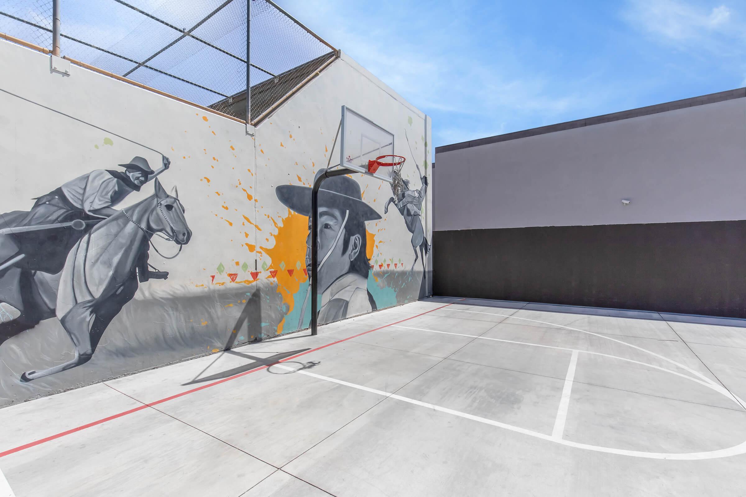 A basketball court featuring a colorful mural on the wall, depicting a horse and rider alongside a stylized figure. The court has a hoop mounted on the wall and is surrounded by a chain-link fence, with a clear blue sky in the background. The ground is marked with court lines.