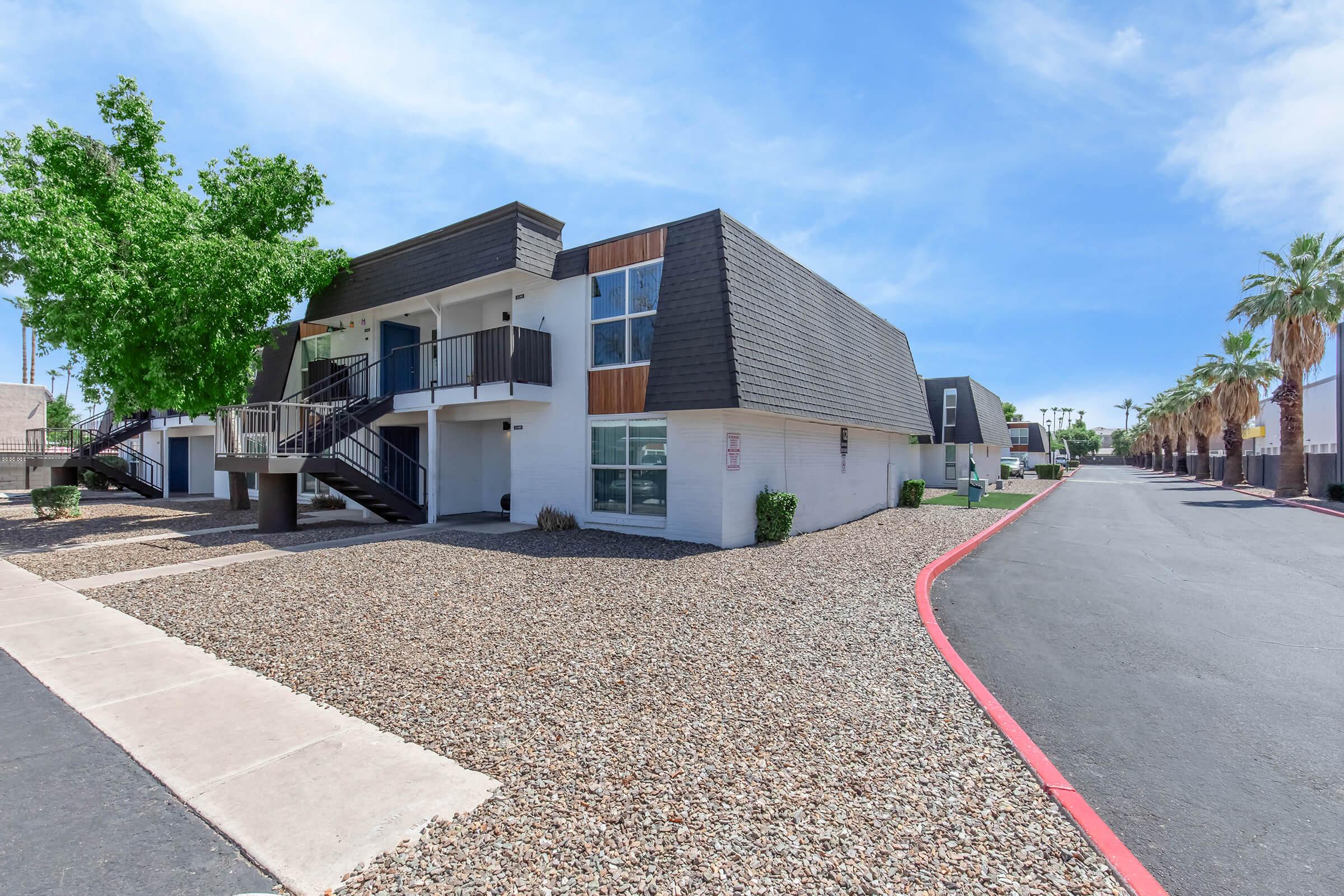 A two-story apartment building with a modern design, featuring a sloped roof and exterior stairs. Surrounding the building is a gravel landscape with palm trees and a paved road in the foreground. The sky is clear and blue, creating a bright atmosphere.