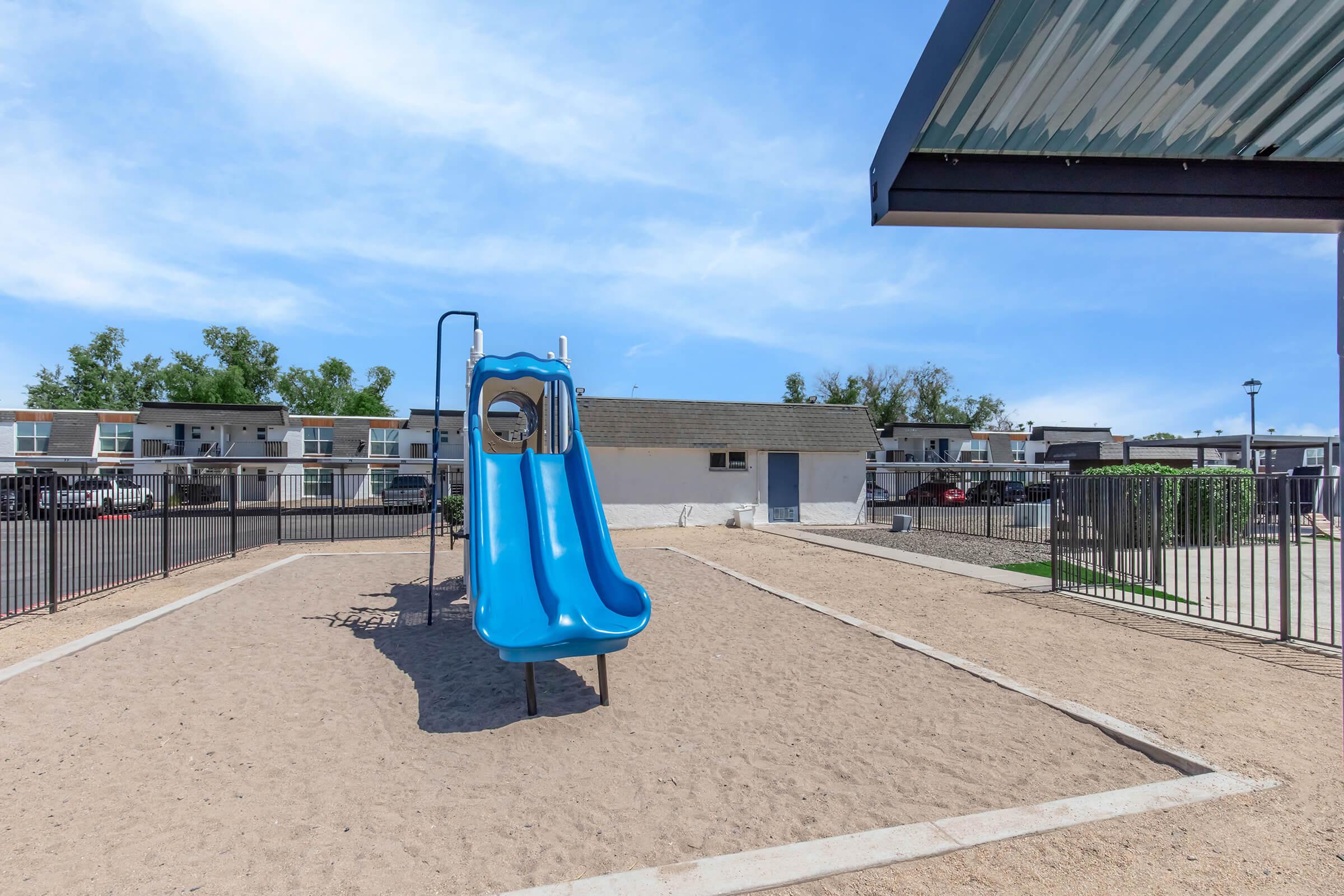 Playground area featuring a blue slide on a sandy surface, surrounded by a black fence. In the background, there are several residential buildings under a clear blue sky. A shaded area with a roof is visible to the right.