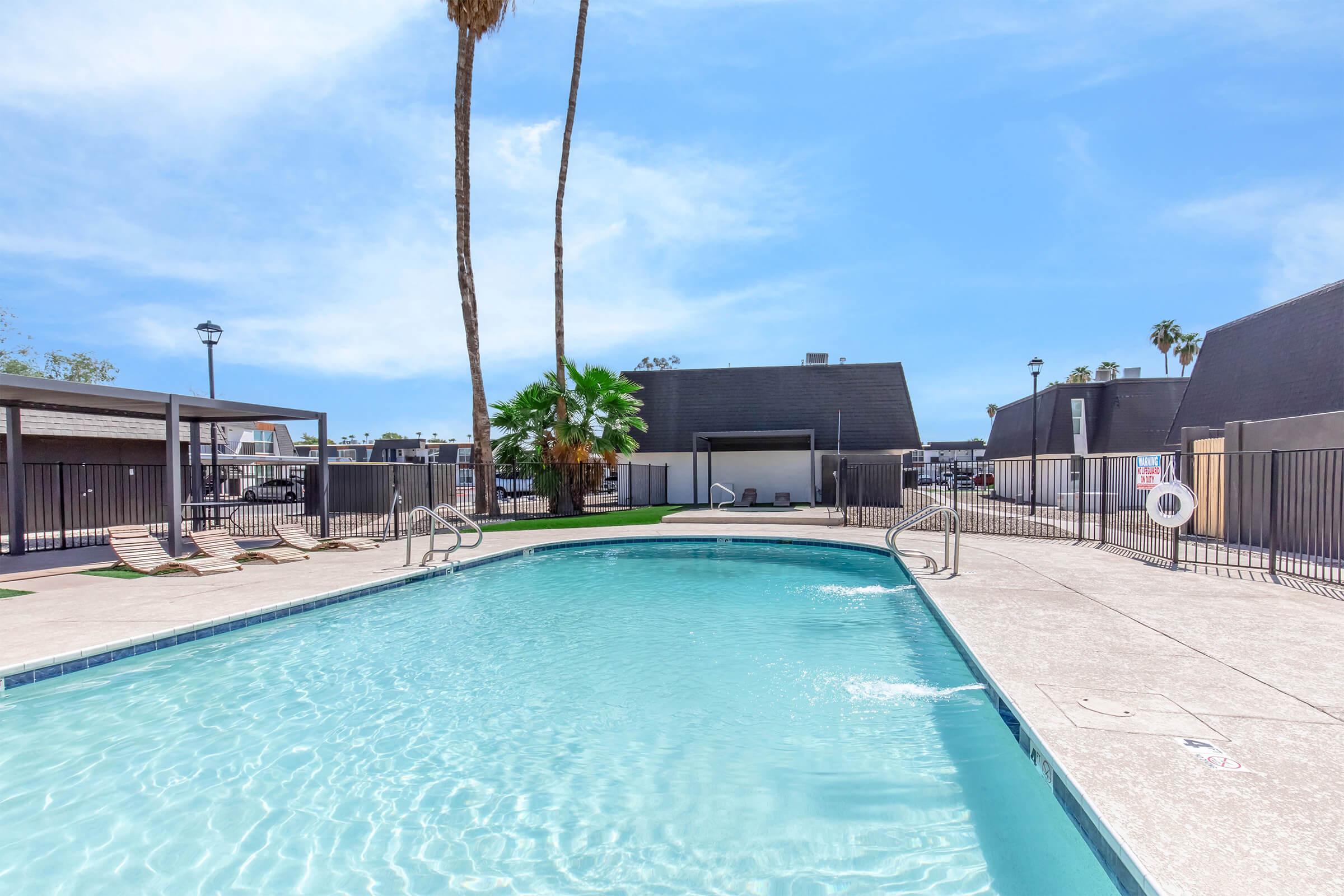 A clear swimming pool surrounded by a concrete deck, with lounge chairs lined up along the edges. Tall palm trees and blue skies in the background create a relaxing atmosphere. Fenced area with shaded cabanas and a few buildings in the distance. Ideal for leisure and recreation.