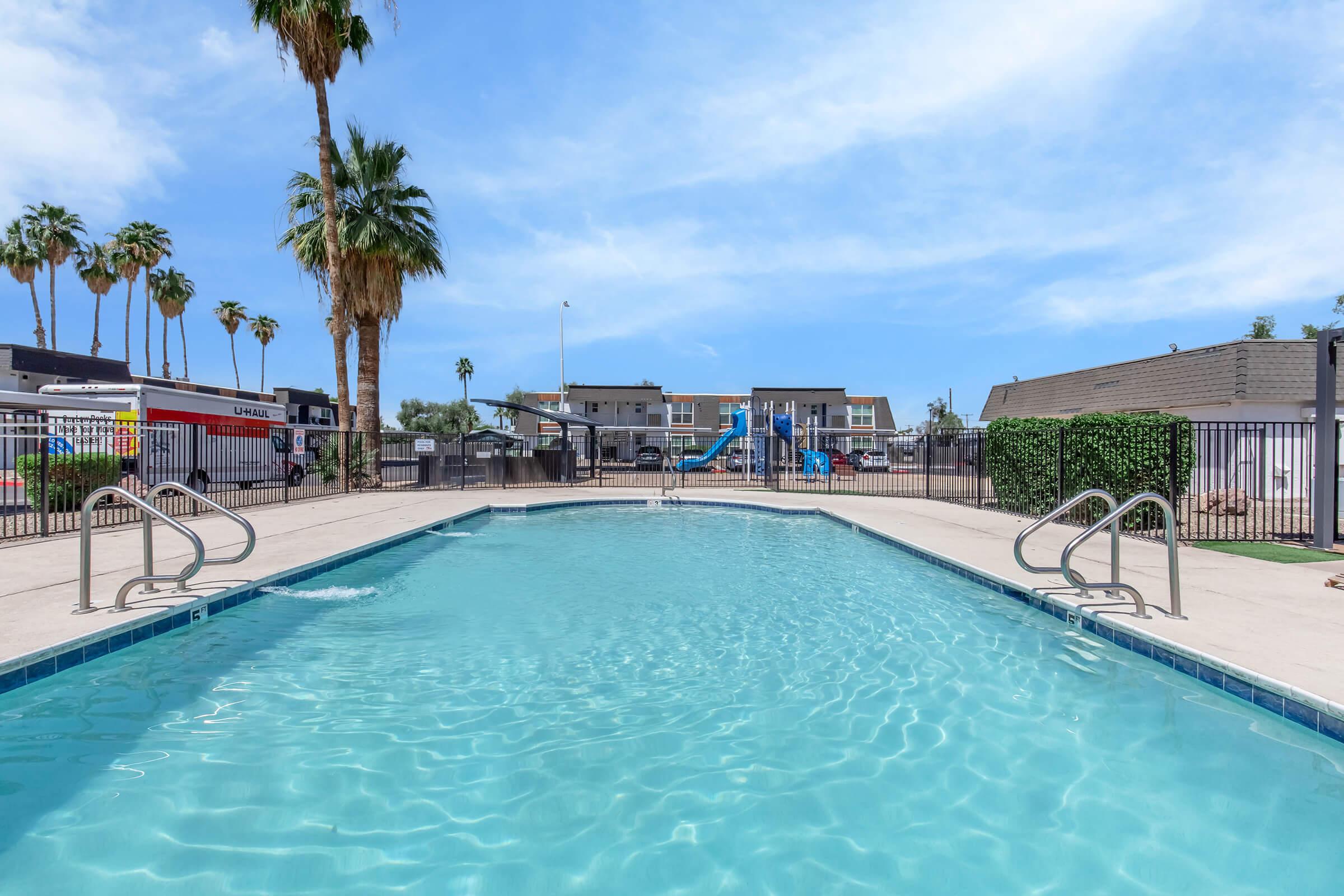 A clear swimming pool surrounded by a concrete deck, with palm trees in the background. A children's slide is visible near the pool, along with lounge chairs. The area is well-lit under a blue sky, creating a welcoming outdoor space.