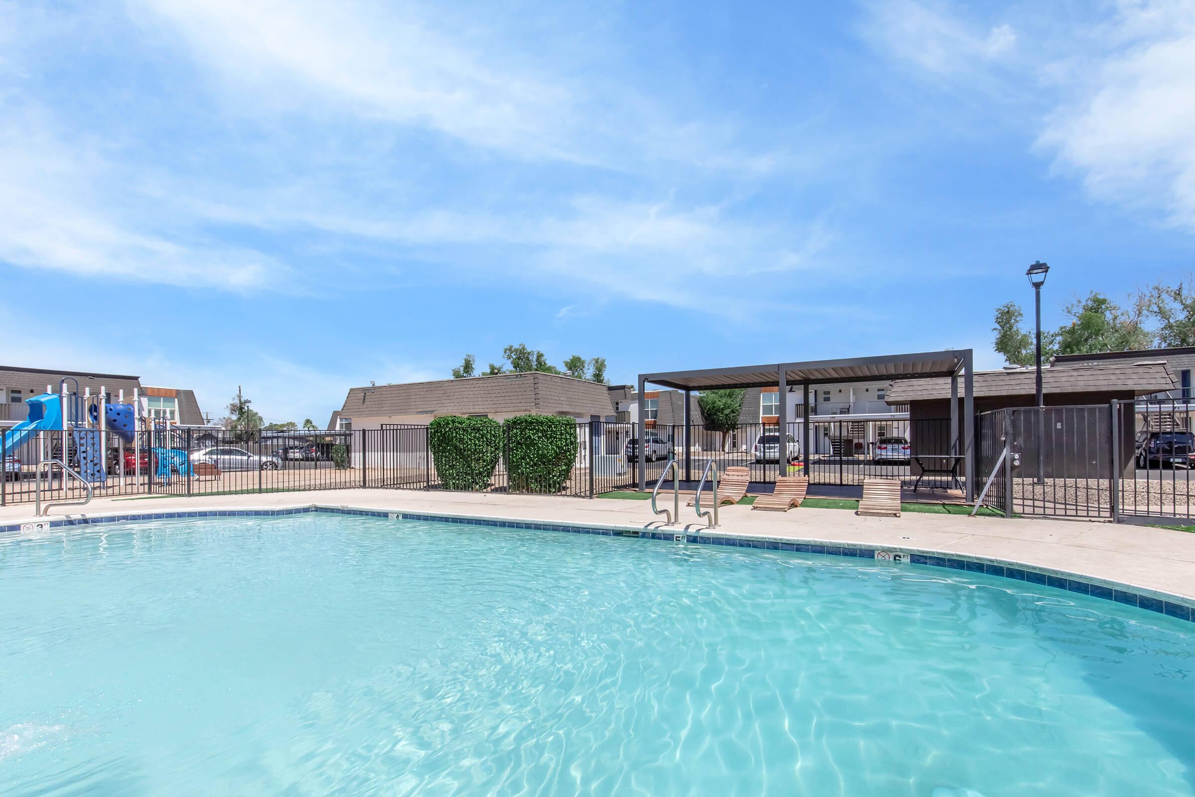 A clear blue swimming pool surrounded by lounge chairs and a shaded area. In the background, there are playground equipment and buildings. The sky is bright with a few clouds, indicating a sunny day. The scene conveys a relaxing outdoor space ideal for leisure activities.