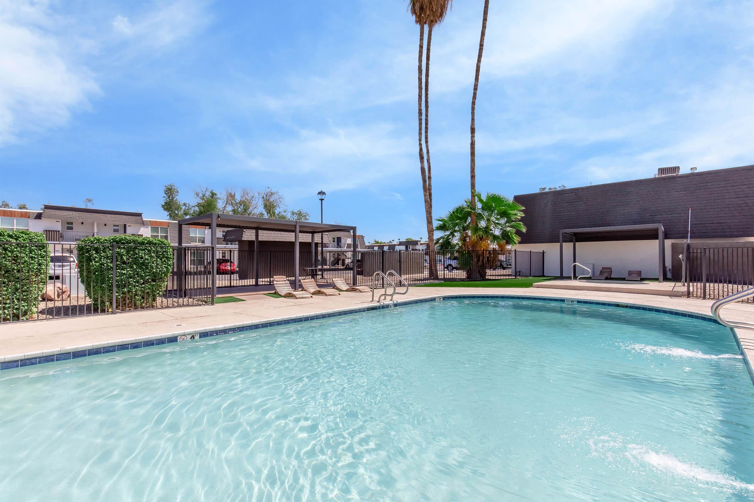 A clear swimming pool surrounded by palm trees and lounge chairs. The area features a fenced-in pool with a sunny blue sky above and nearby buildings in the background. The setting is tranquil, ideal for relaxation and leisure.