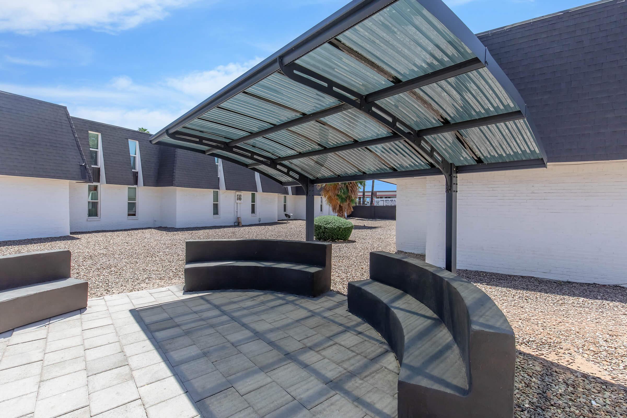 A shaded seating area featuring a curved, dark bench beneath a clear corrugated roof. The surrounding landscape consists of gravel and low shrubs, with white-walled buildings in the background under a blue sky.