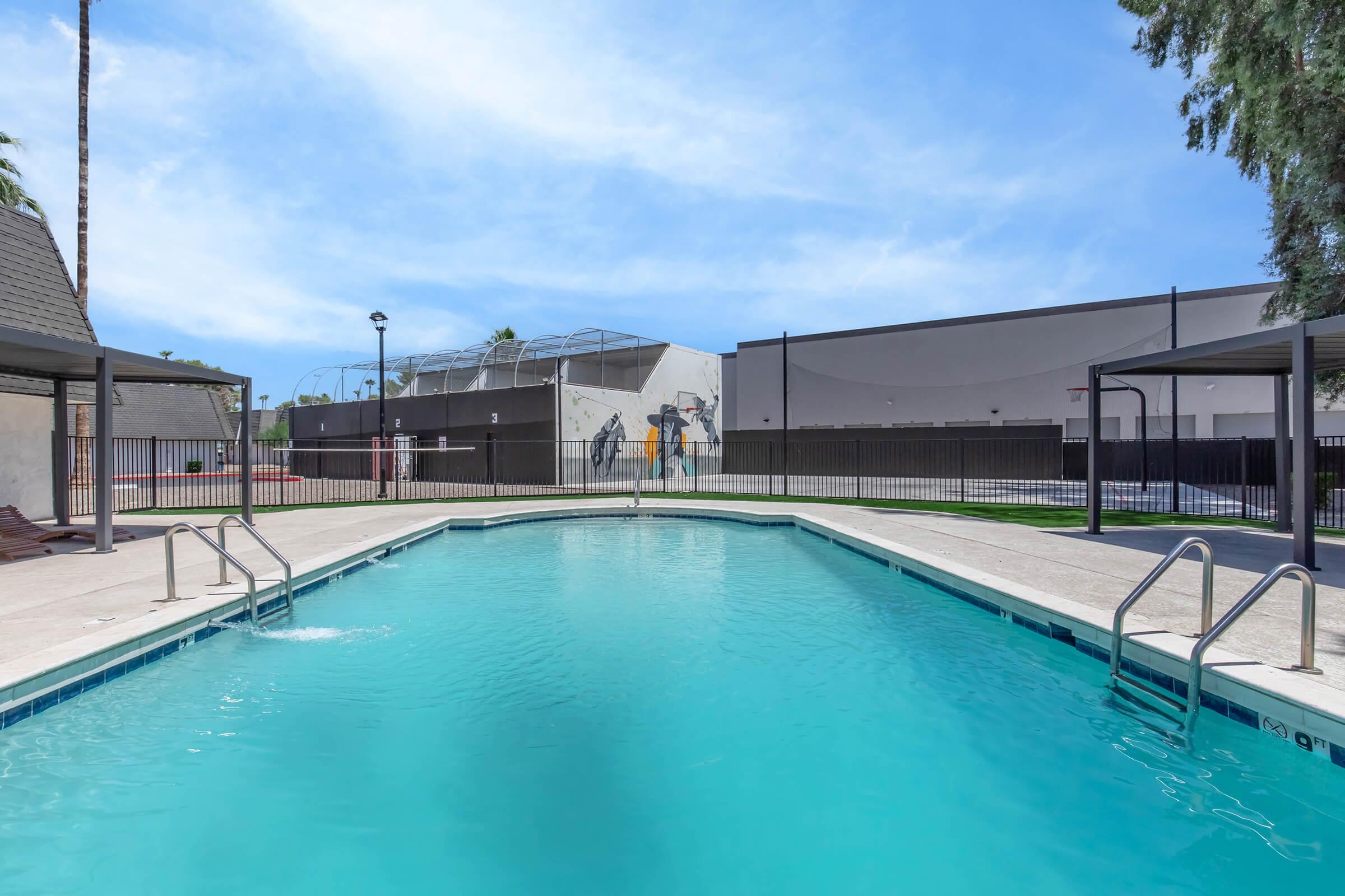 A clear swimming pool with a diving ledge, surrounded by concrete decking. In the background, there are basketball courts and a large building. Palm trees are visible nearby, and the sky is bright with a few clouds. The scene is inviting and well-maintained, ideal for relaxation and recreation.
