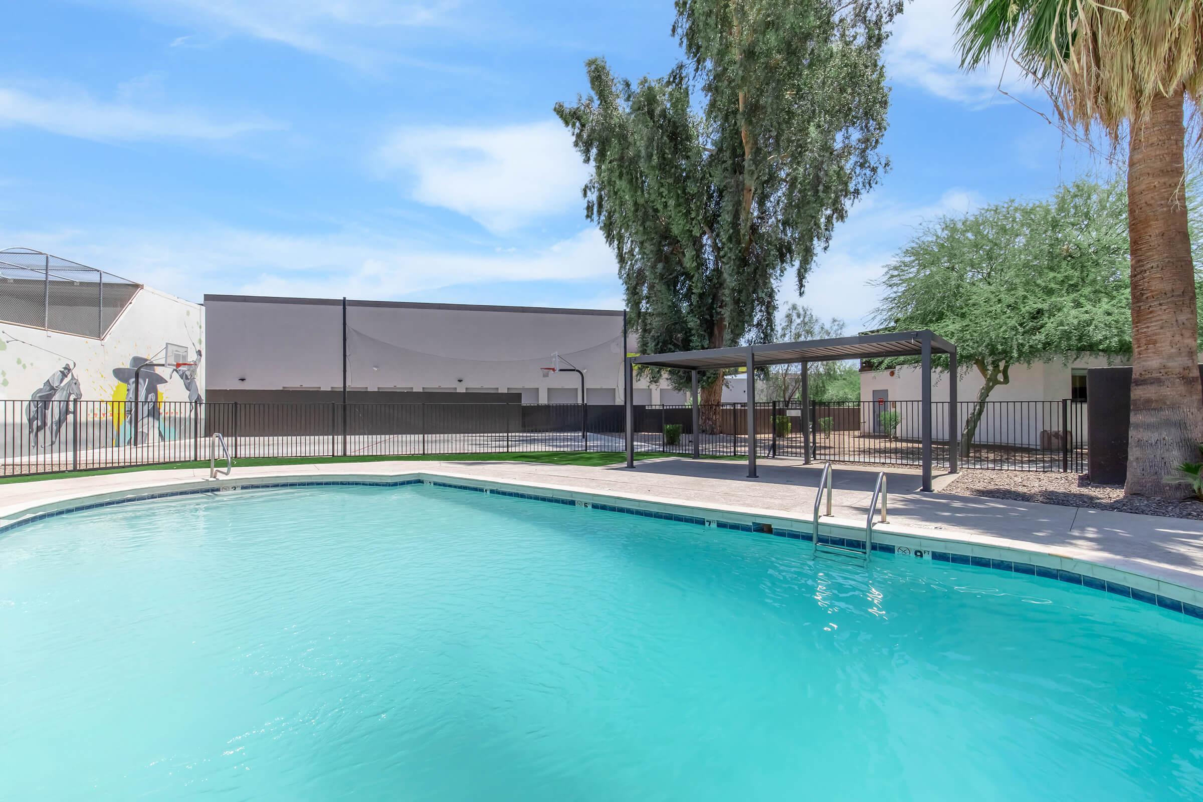 A serene outdoor swimming pool surrounded by a black metal fence. There are palm trees nearby, and a shaded seating area with a gazebo is visible. In the background, a wall and basketball hoop can be seen. The sky is clear with a few clouds.