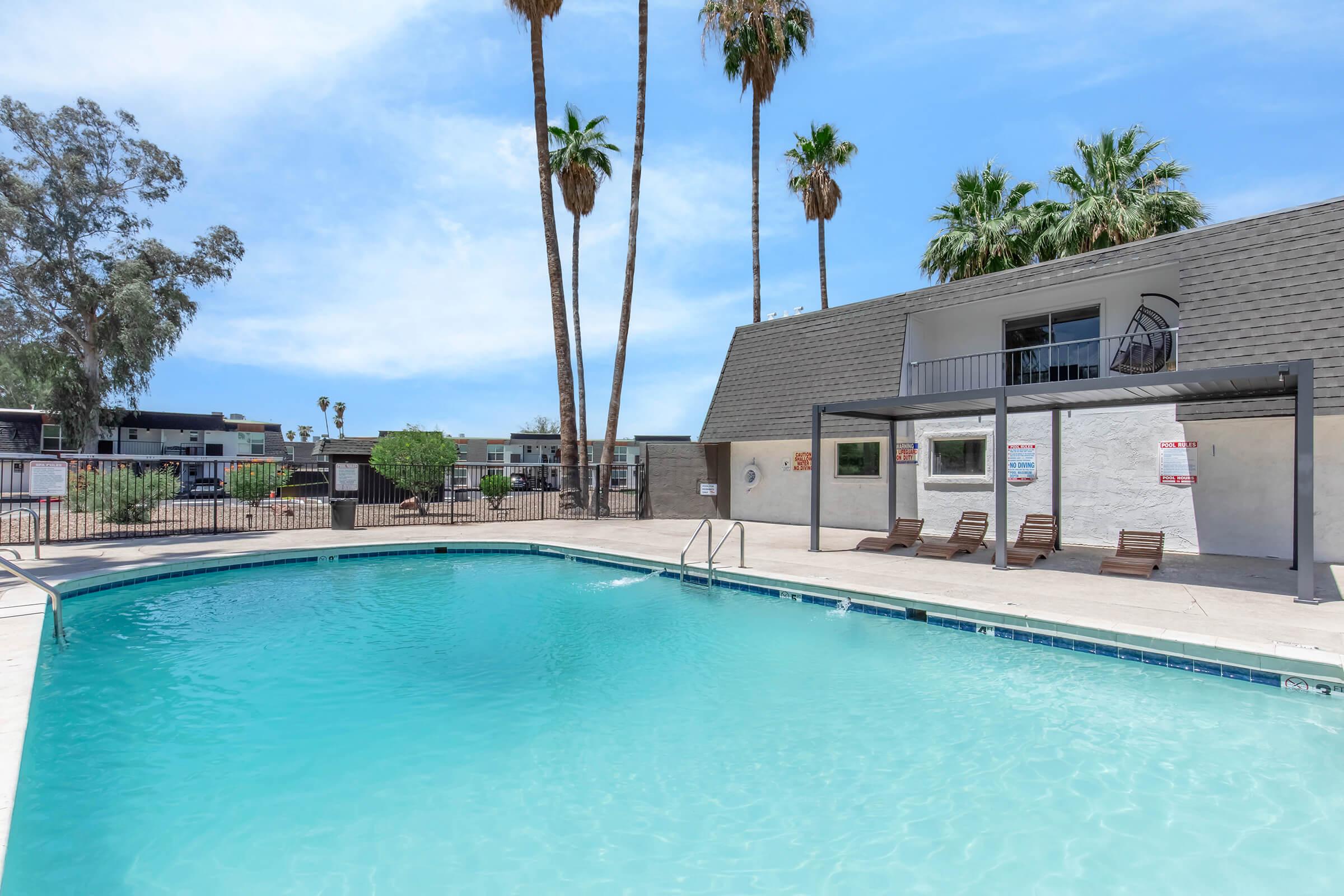 A clear swimming pool surrounded by palm trees and a sunbathing area with lounge chairs. In the background, there are apartment buildings and a blue sky. The setting suggests a serene and inviting atmosphere for relaxation and leisure.