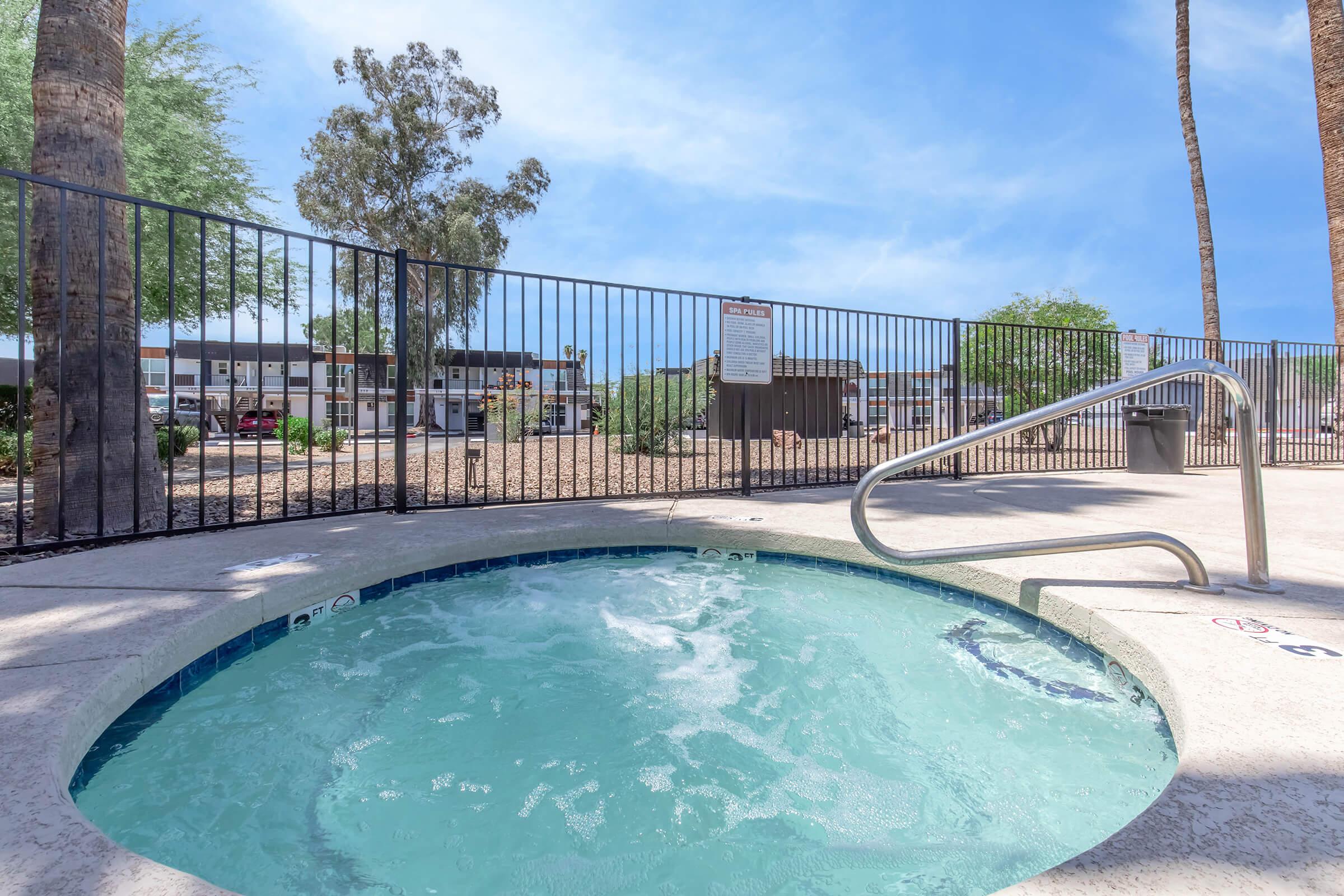 A hot tub with bubbling water, surrounded by a fenced area. In the background, there are palm trees and a clear blue sky. The setting appears to be part of a residential complex, with visible signs and landscaping enhancing the outdoor space.